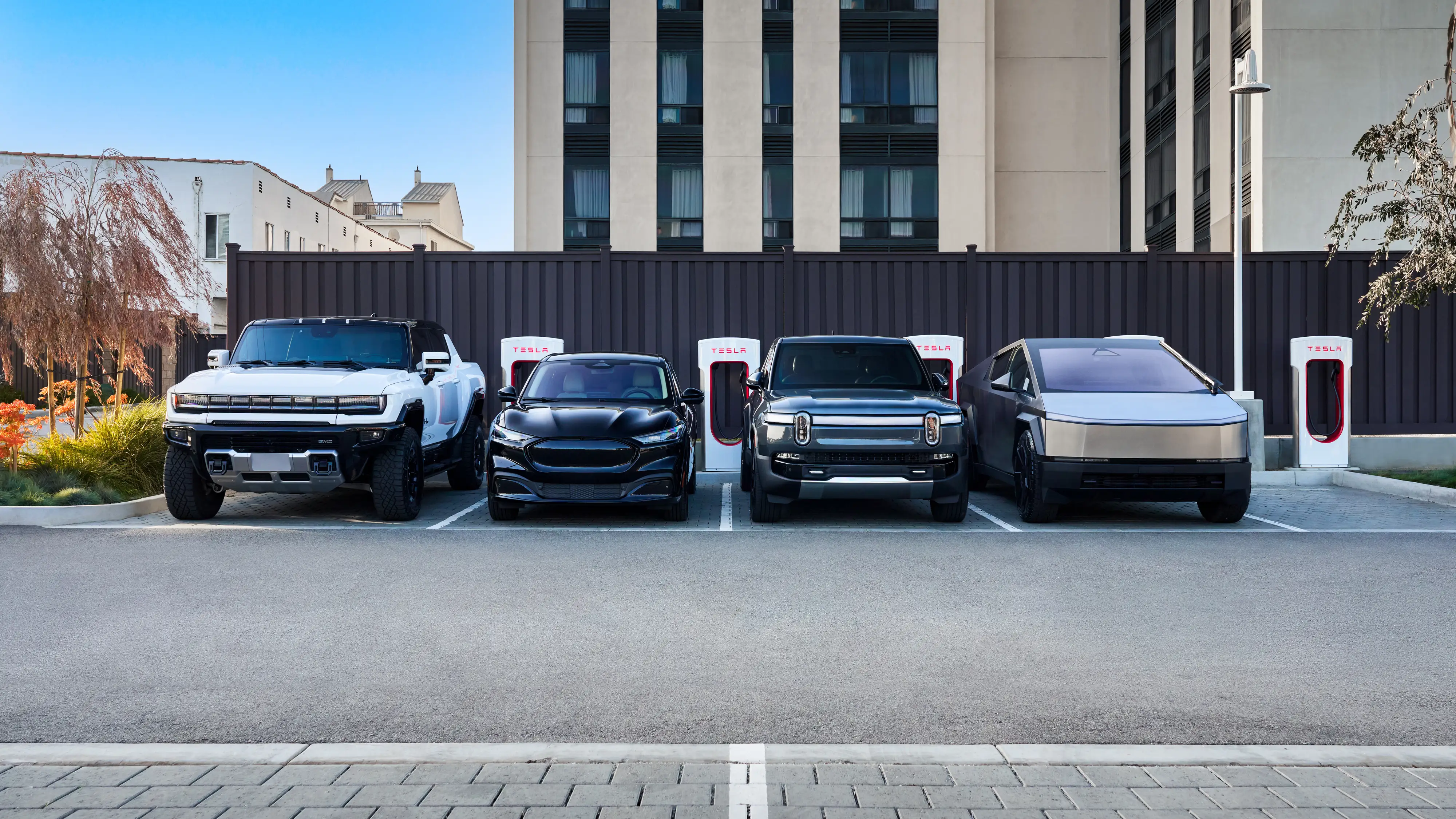 A line of electric vehicles — including a GMC, Ford, Rivian, and Tesla — is parked next to a row of Tesla Superchargers.