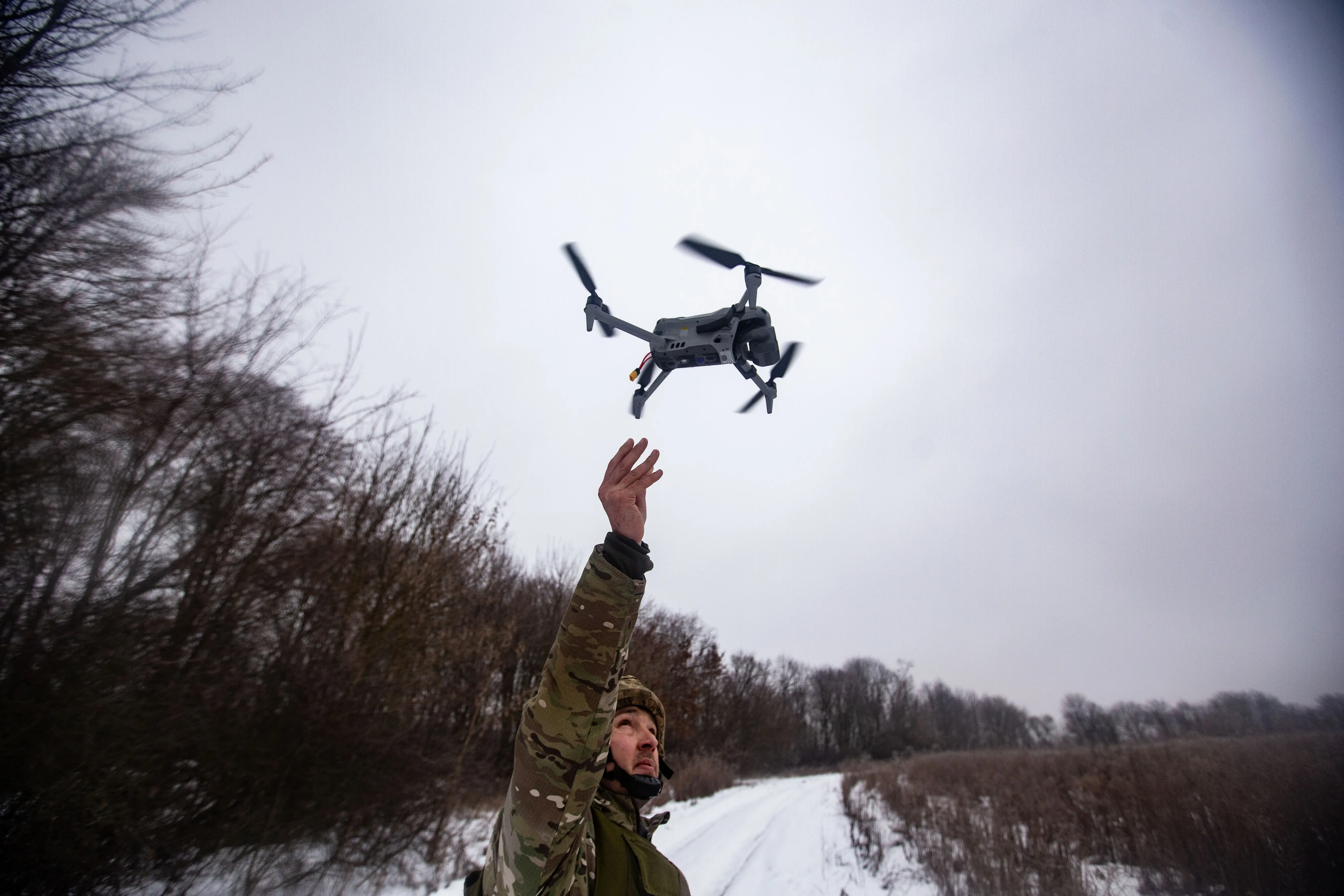 A man in camouflage stands on a snowy road between trees with his arm outstretched to a small grey drone in a grey sky