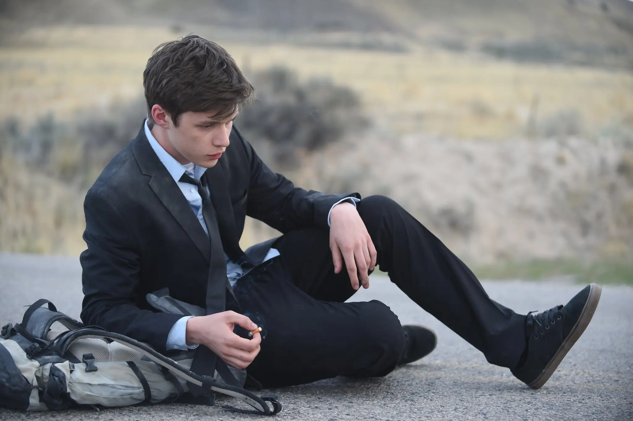 Nick Robinson in a suit and tie sitting on the ground in Being Charlie movie