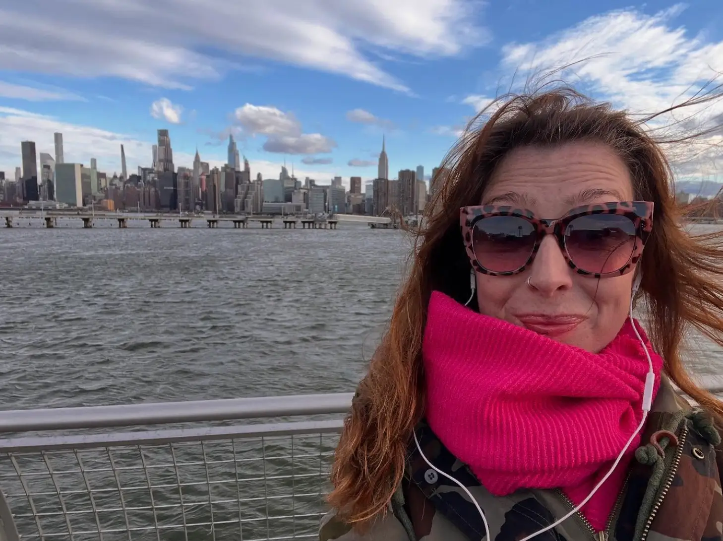 The writer wearing sunglasses and a scarf, posing in front of the water with New York buildings in the background.