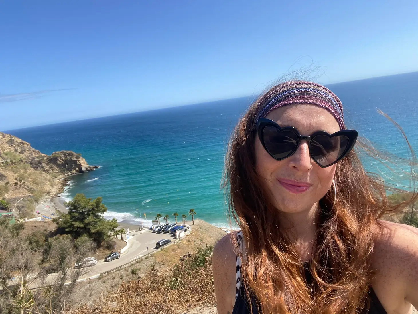 The writer wearing heart-shaped sunglasses and posing in front of blue water in Spain.