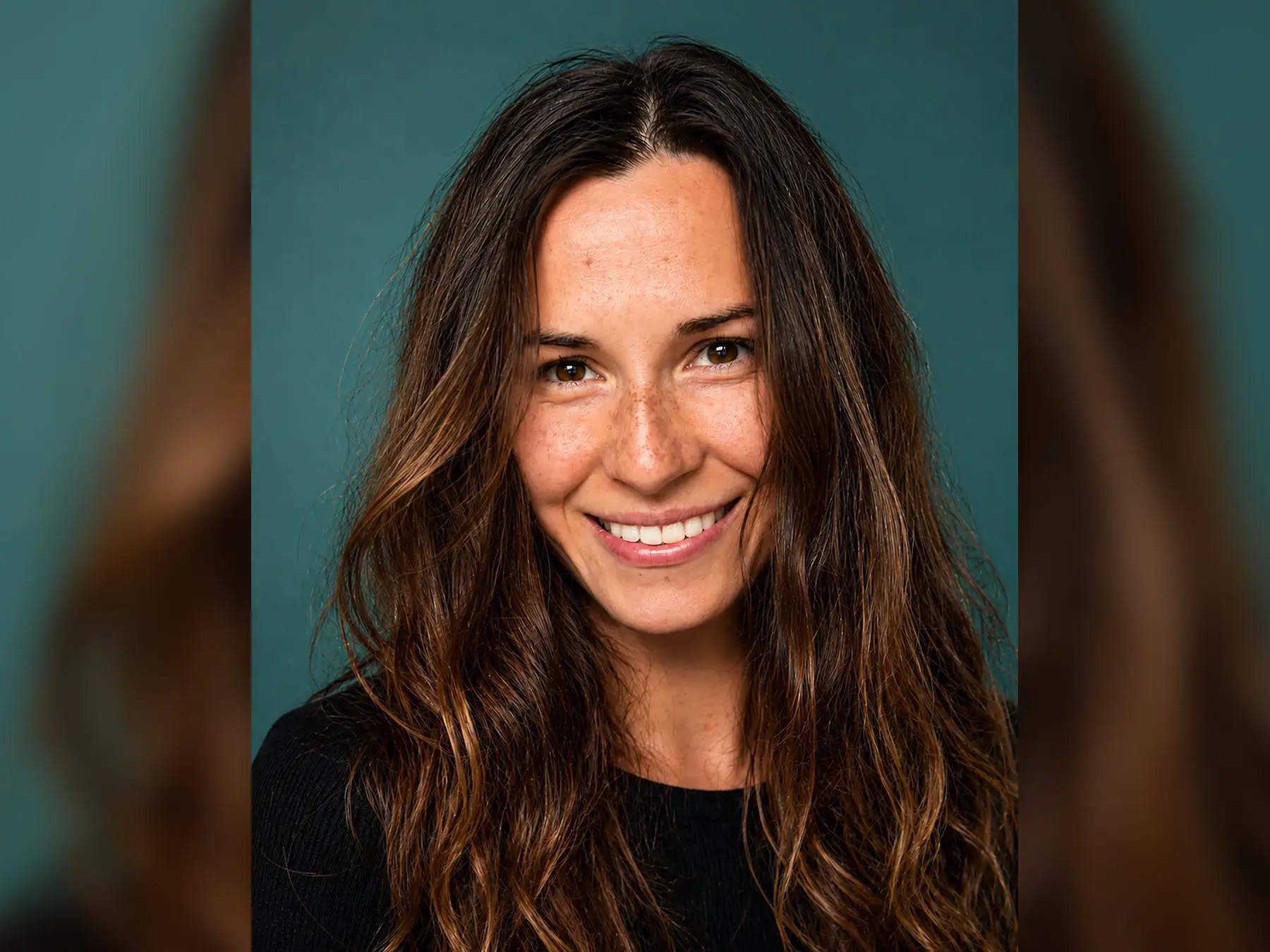 A smiling woman with long brown hair poses against a teal background.