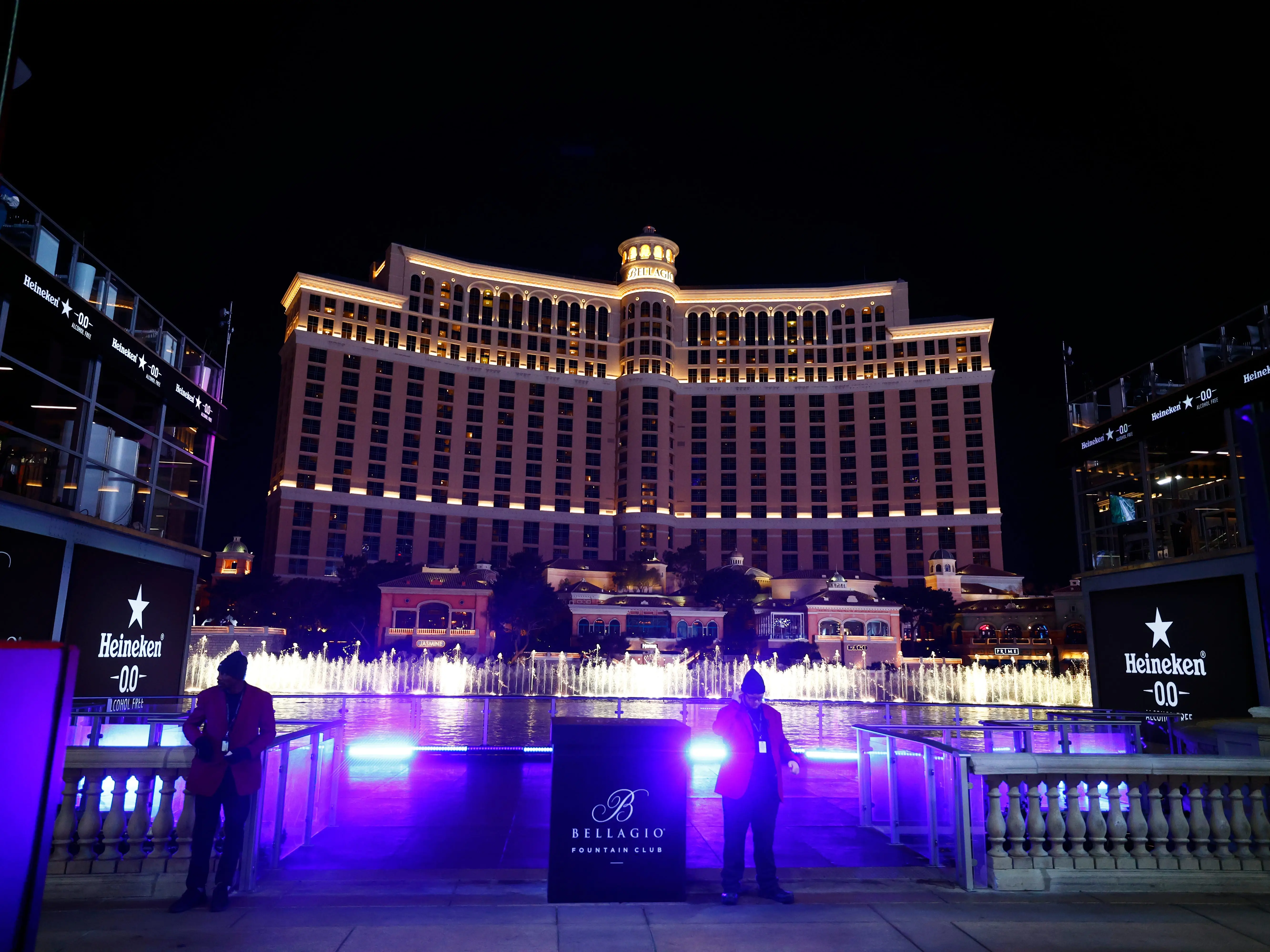 A large building with many windows and fountains glowing blue out front with people looking at it behind a fence