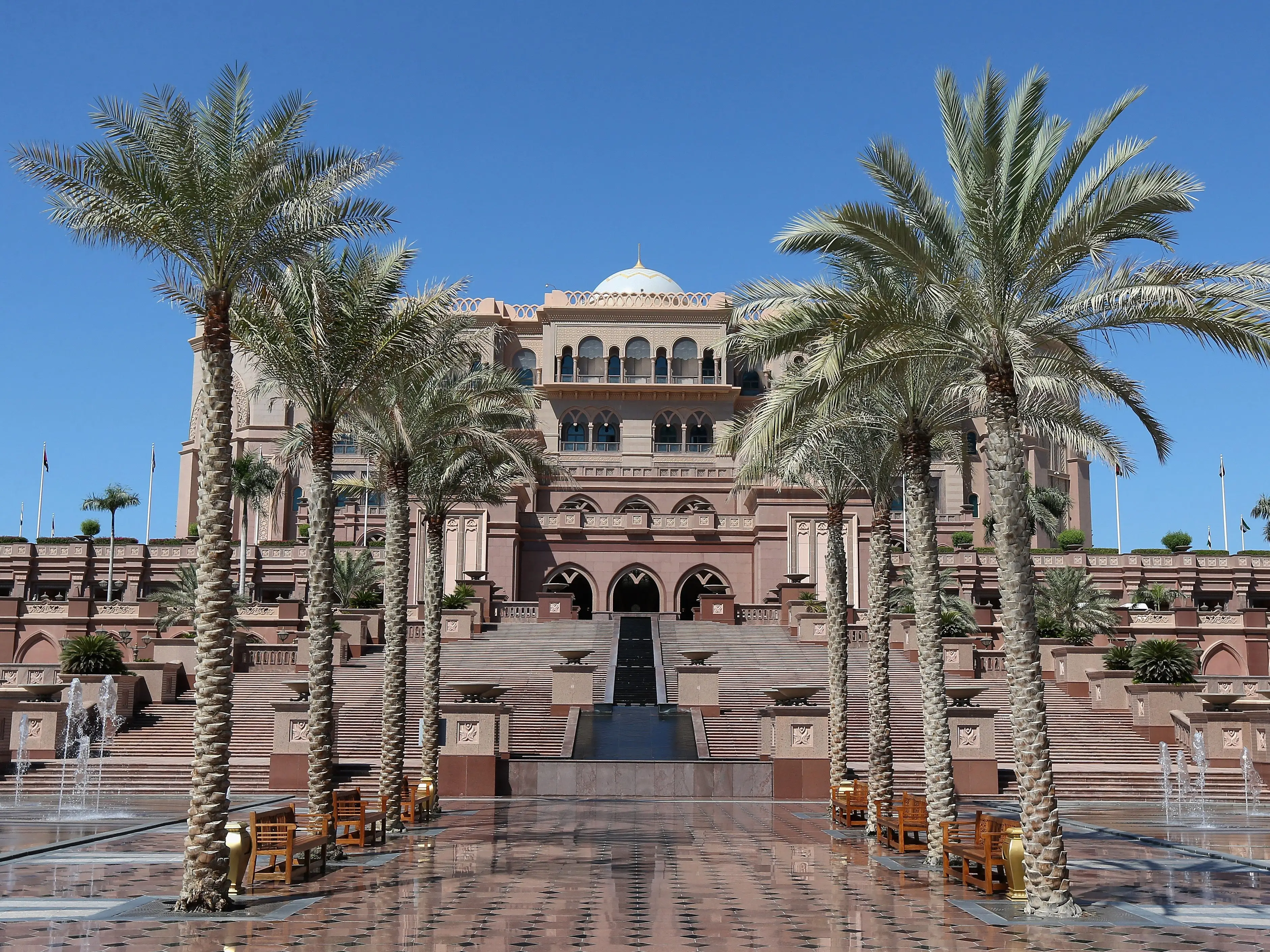 Palm trees and fountains in front of a large sand-colored building with a dome on top