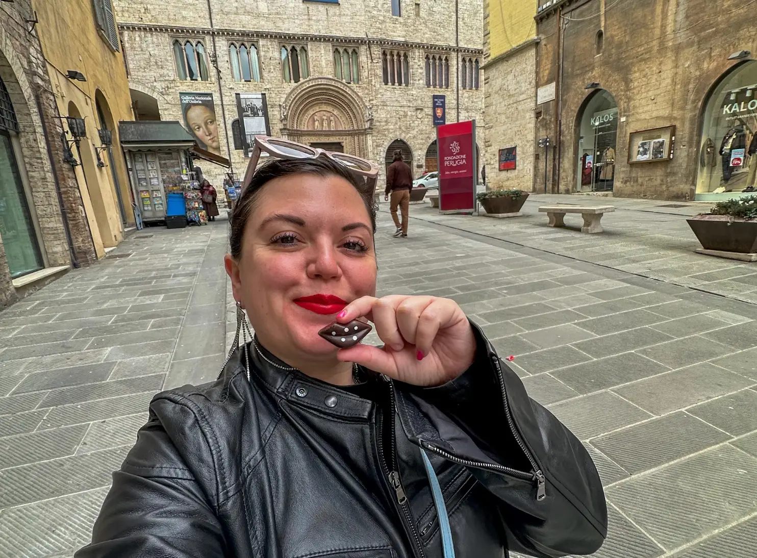 The writer wearing a leather jacket, holding a chocolate, and posing in front of an old museum in Perugia.