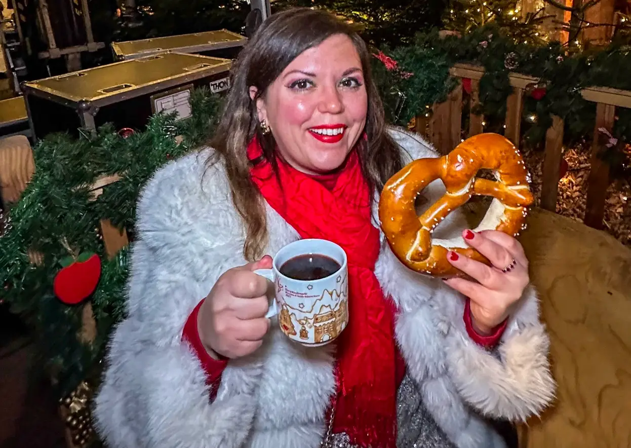 The writer wearing a fur coat, drinking something from a mug, and holding a massive pretzel in Bolzano.