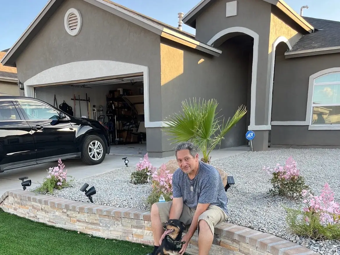 A man sits in front of a house, holding a small dog.