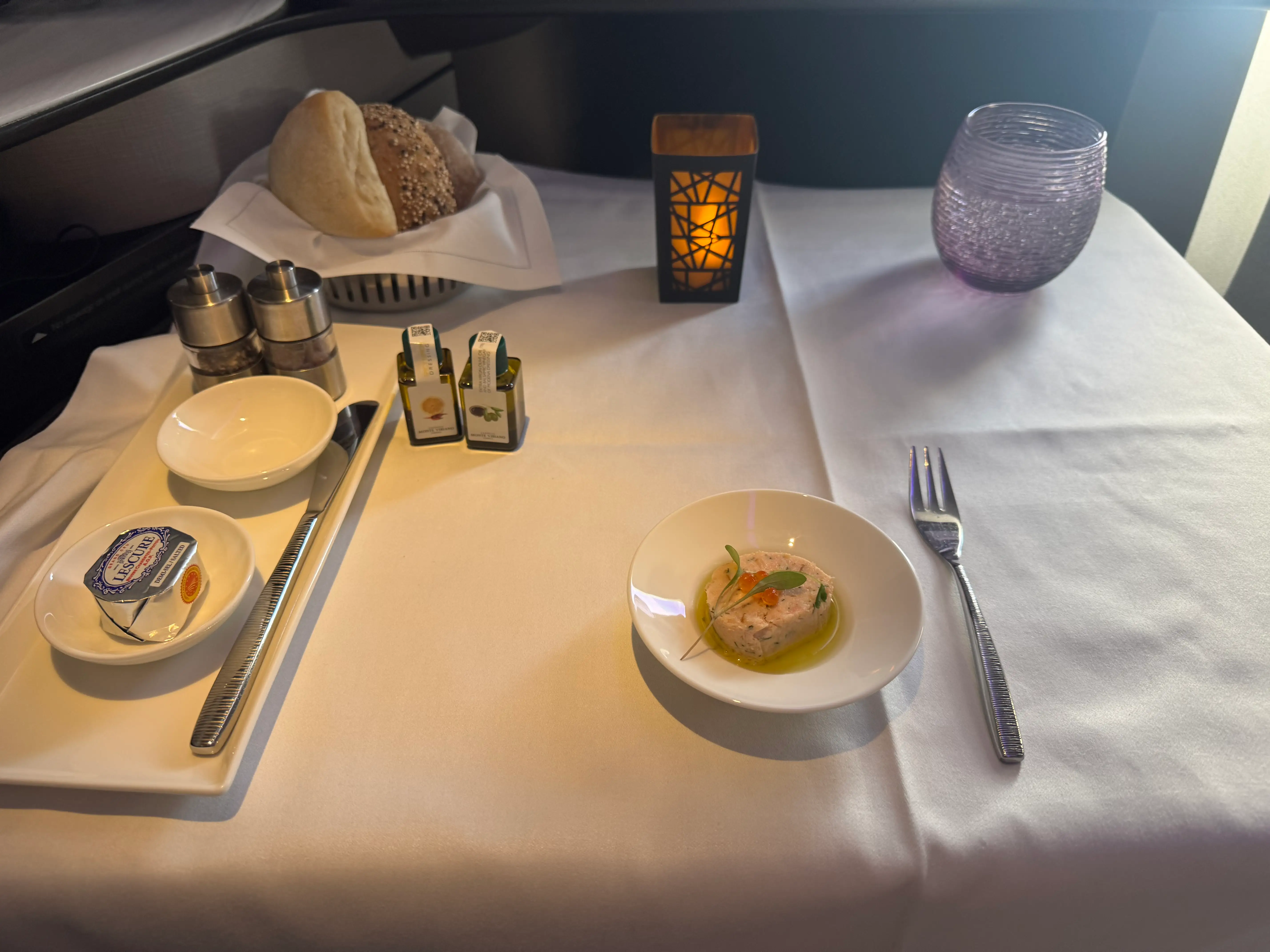 faux candle, bread basket, plated food, silverware on white tablecloth covered tray table on Q Suite flight