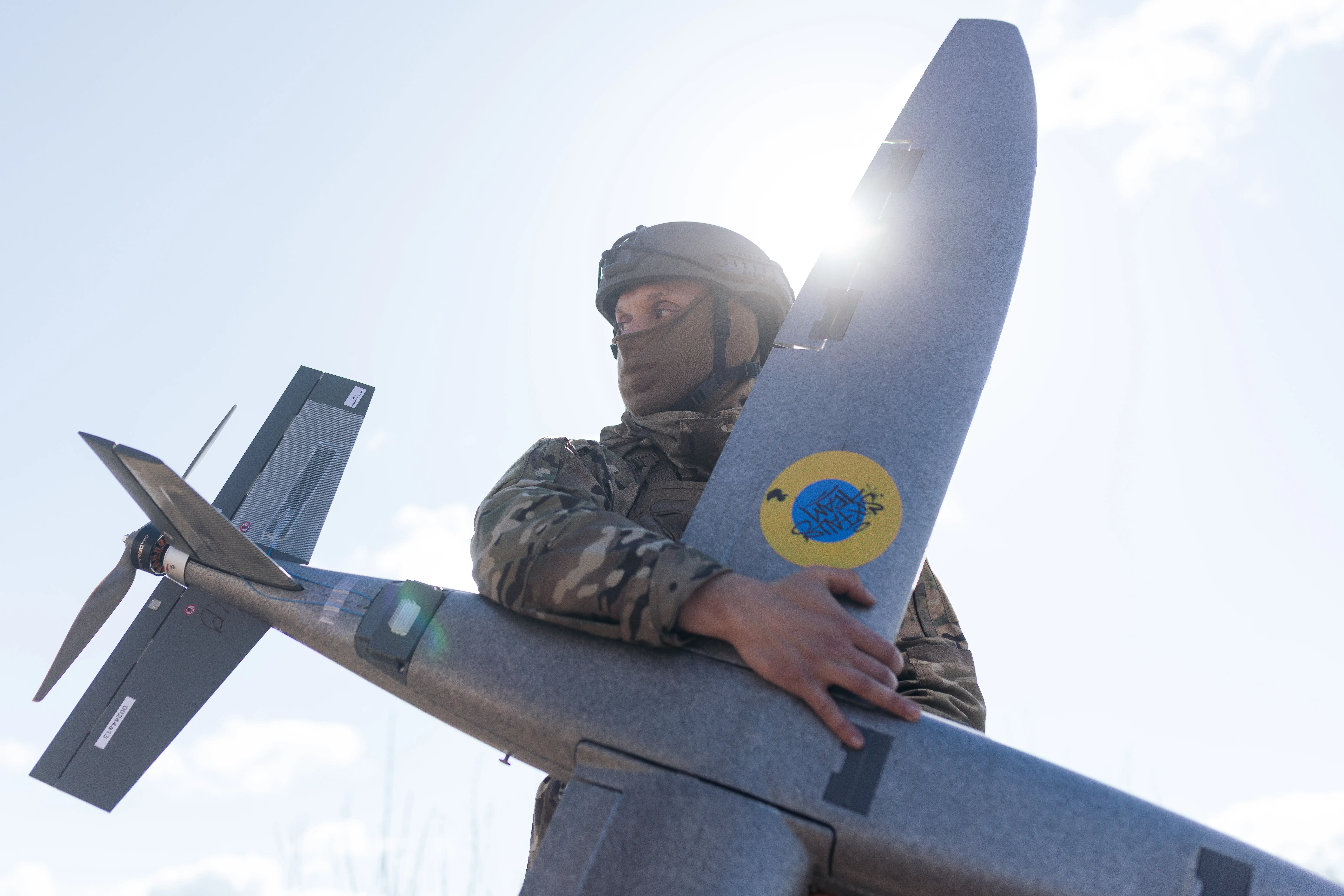 A man in camouflage gear and a helmet stands outside with the sun behind him holding a large grey drone with a blue and yellow circle on it.