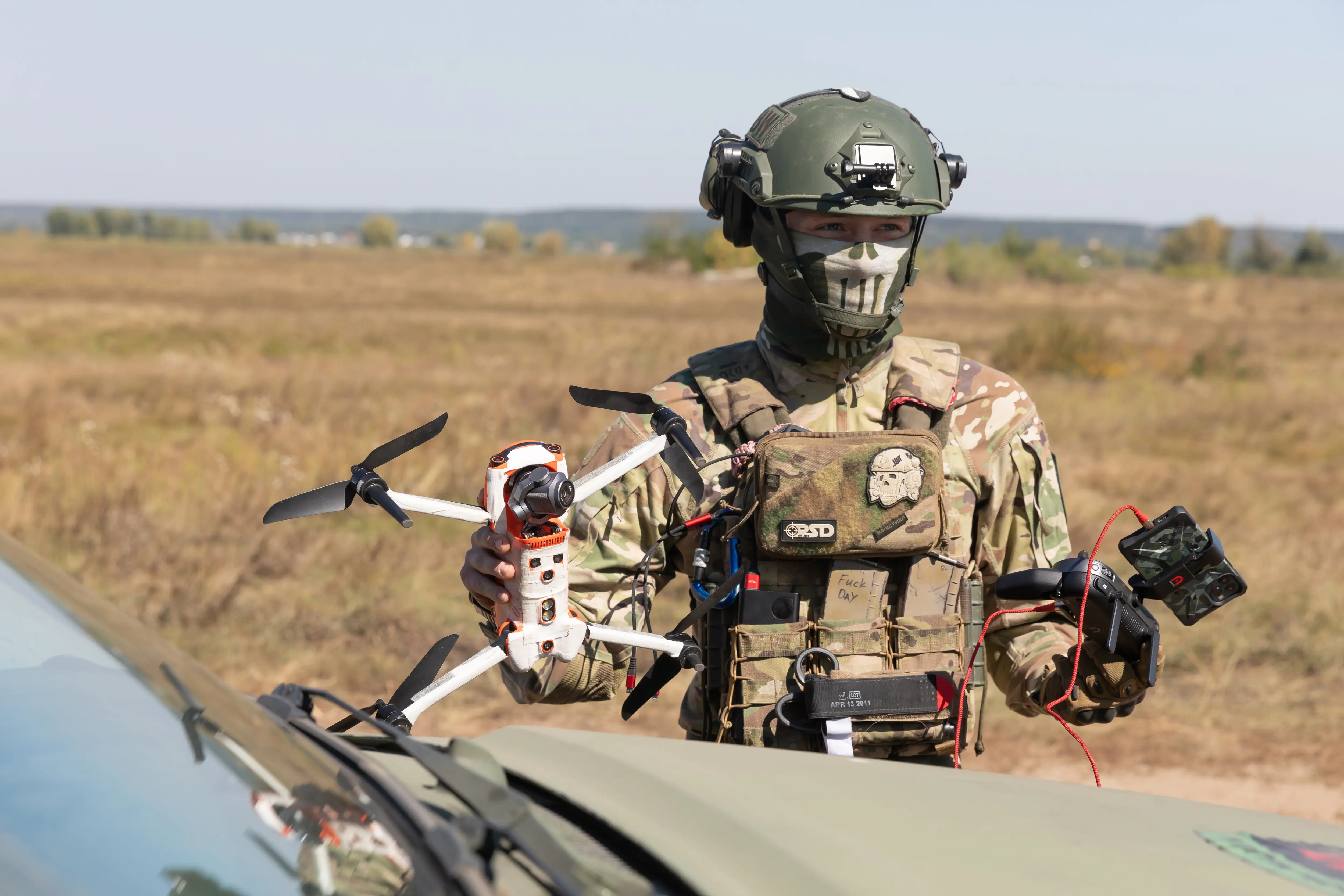A man in camouflage and a helmet stands holding a small white drone in a brown field