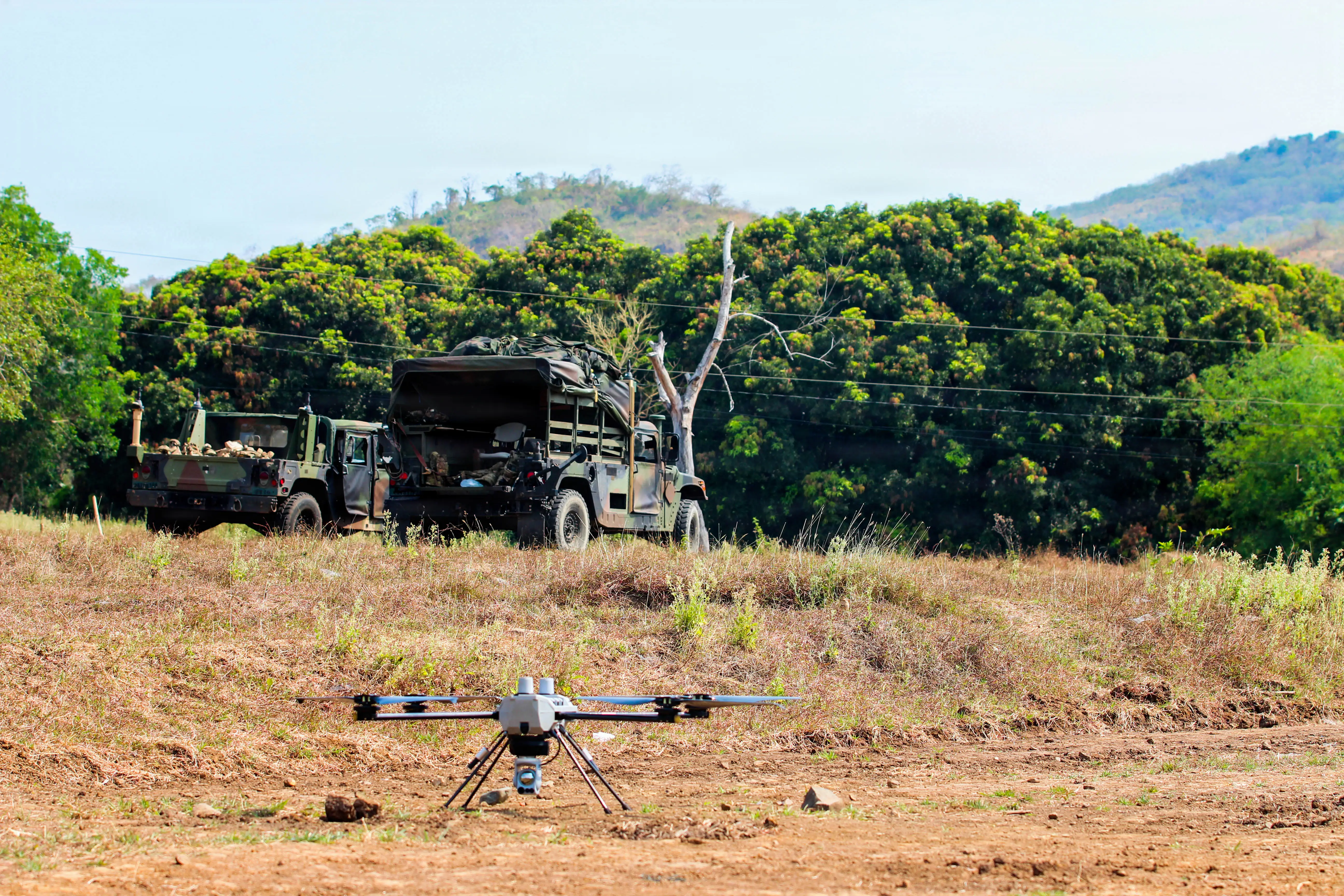 A small drone sits in the dirt in the jungle with vehicles in the distance.