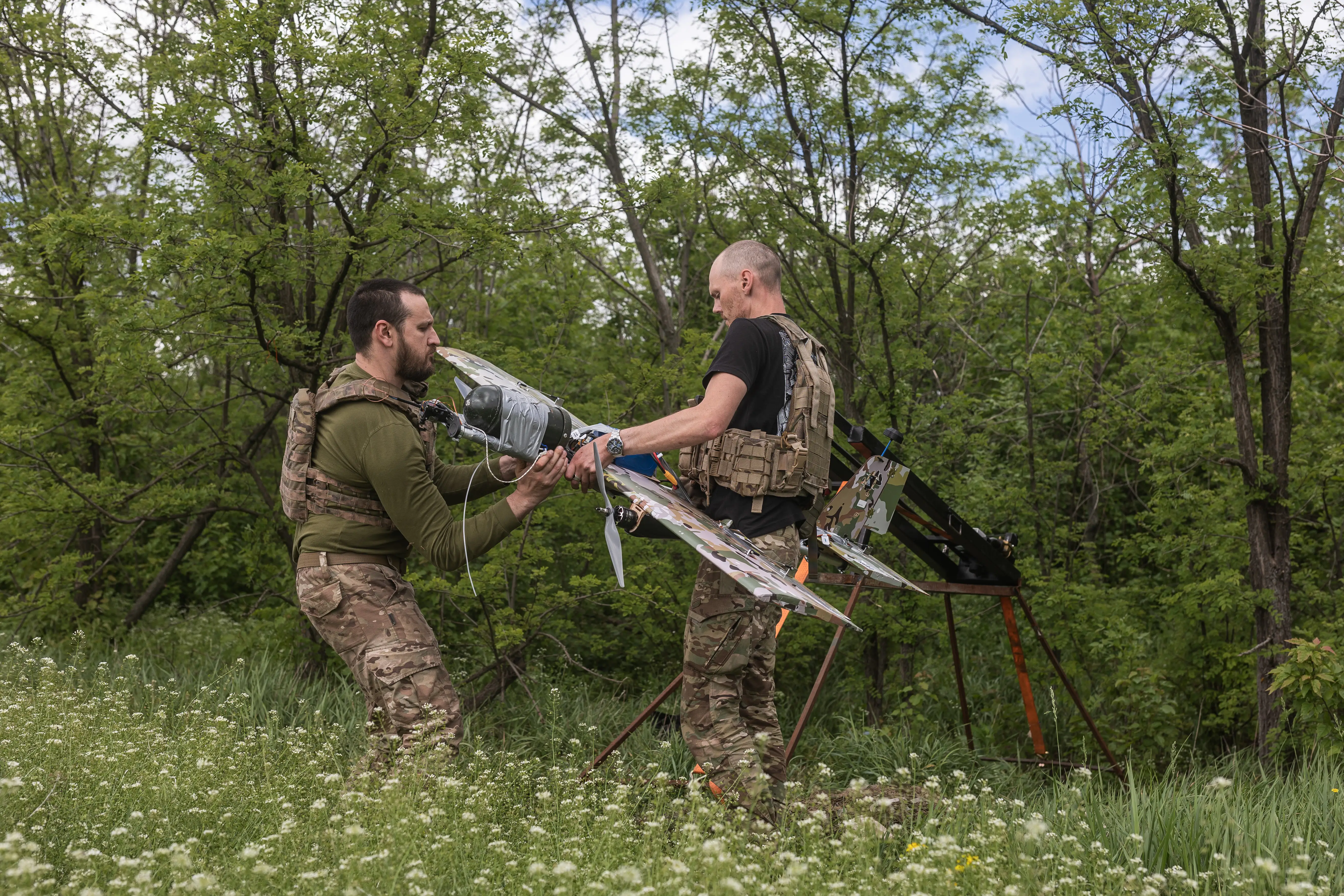 Two men in camouflage hold a black and grey drone in a field with white flowers and trees