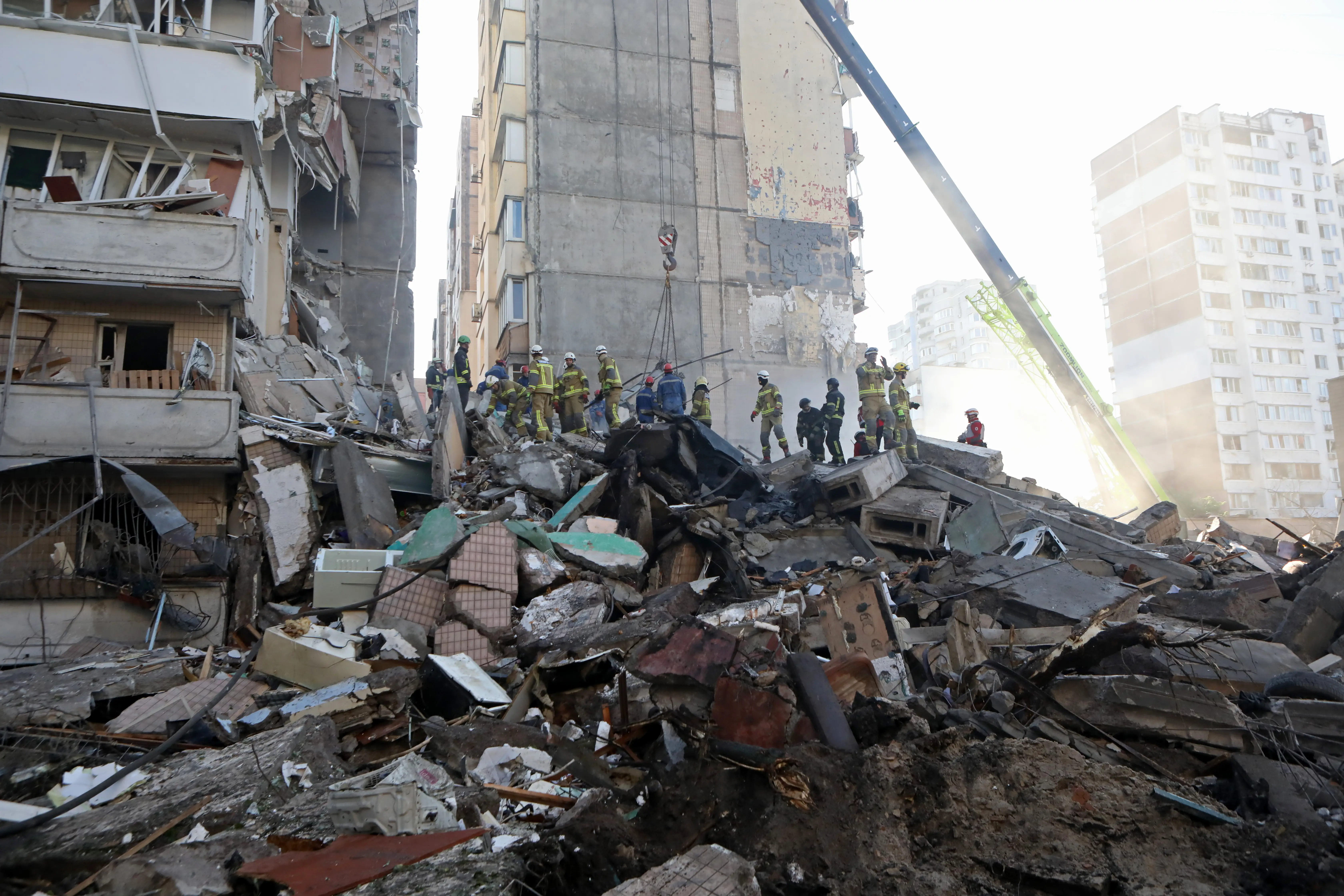 A large pile of rubble among damaged buildings with people in high-vis clothing standing on top of it