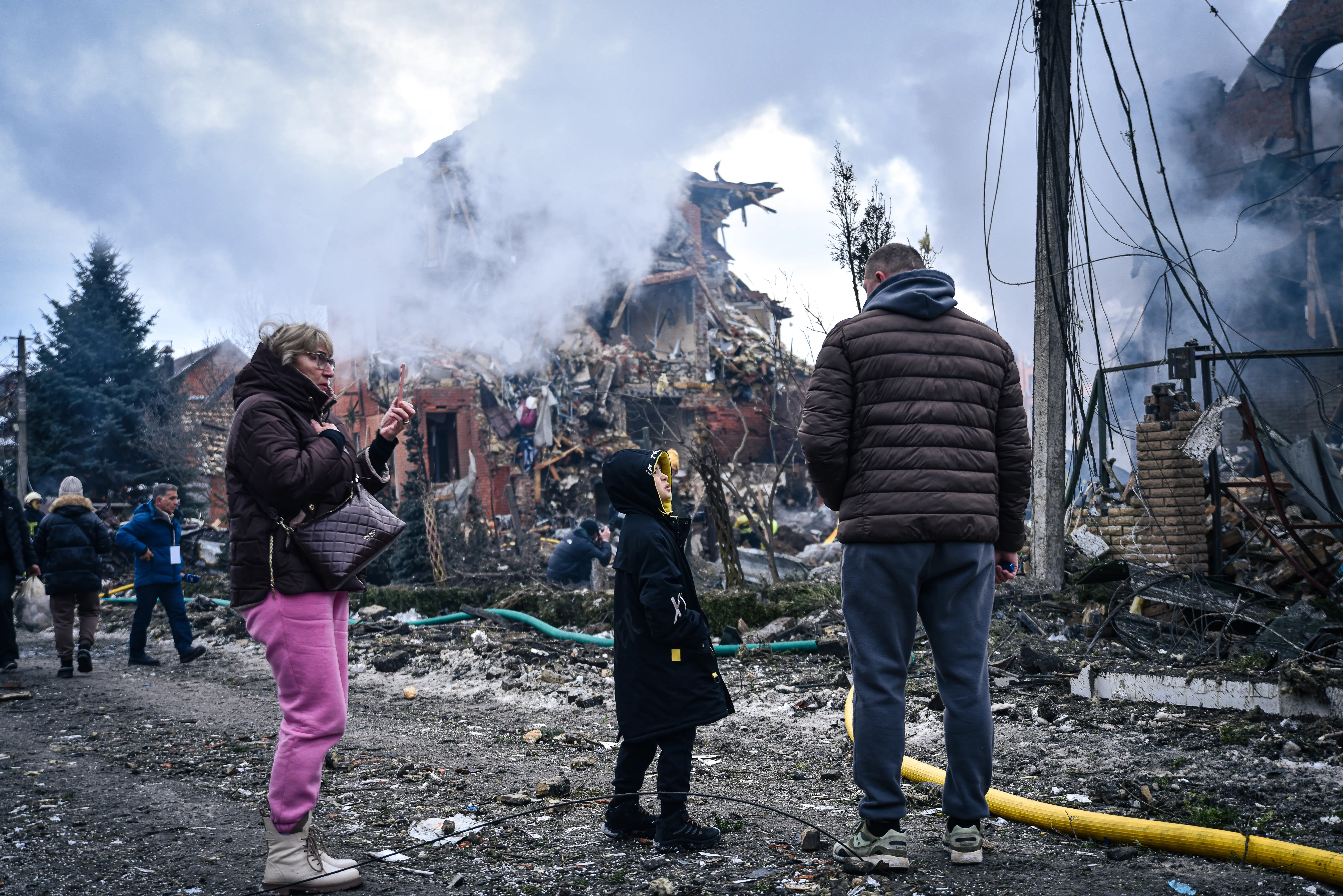 A woman, child, and man stand with thier backs to the camera looking at a destroyed and smoking building