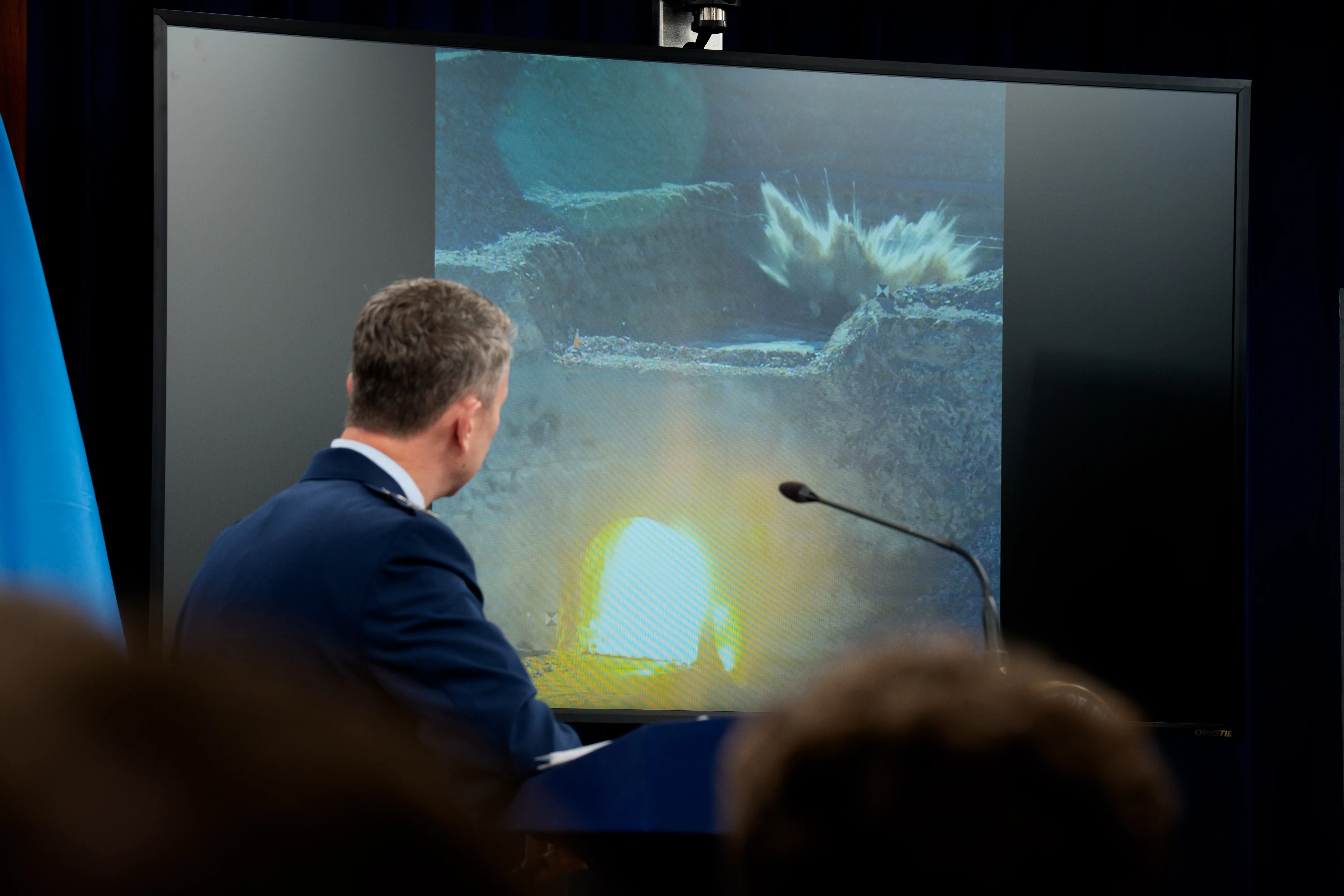 A man stands in front of a screen showing a picture of an explosion.