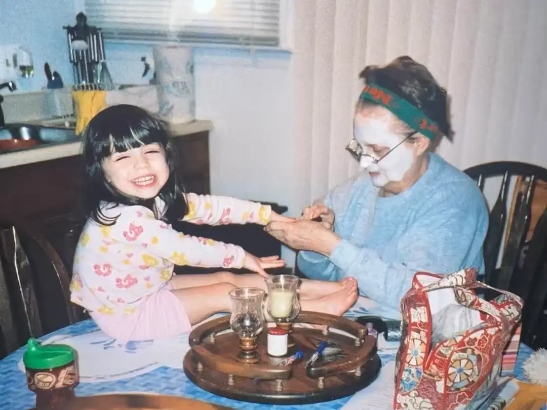 A photo of Amanda as a child, sitting on a table while her grandmother (who is wearing a face mask) paints her nails.