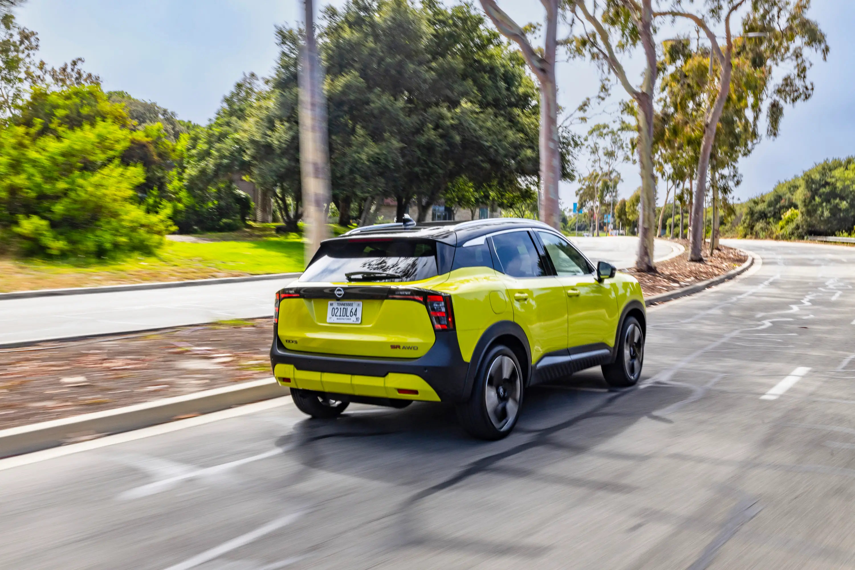 A lime green Nissan Kicks drives down a four-lane road with a tree-lined median.