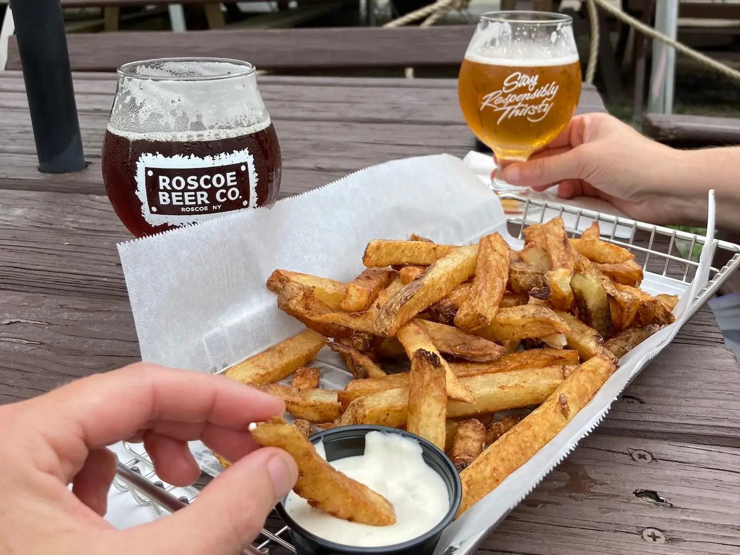 A basket of french fries with dipping sauce and two beers on a picnic table.