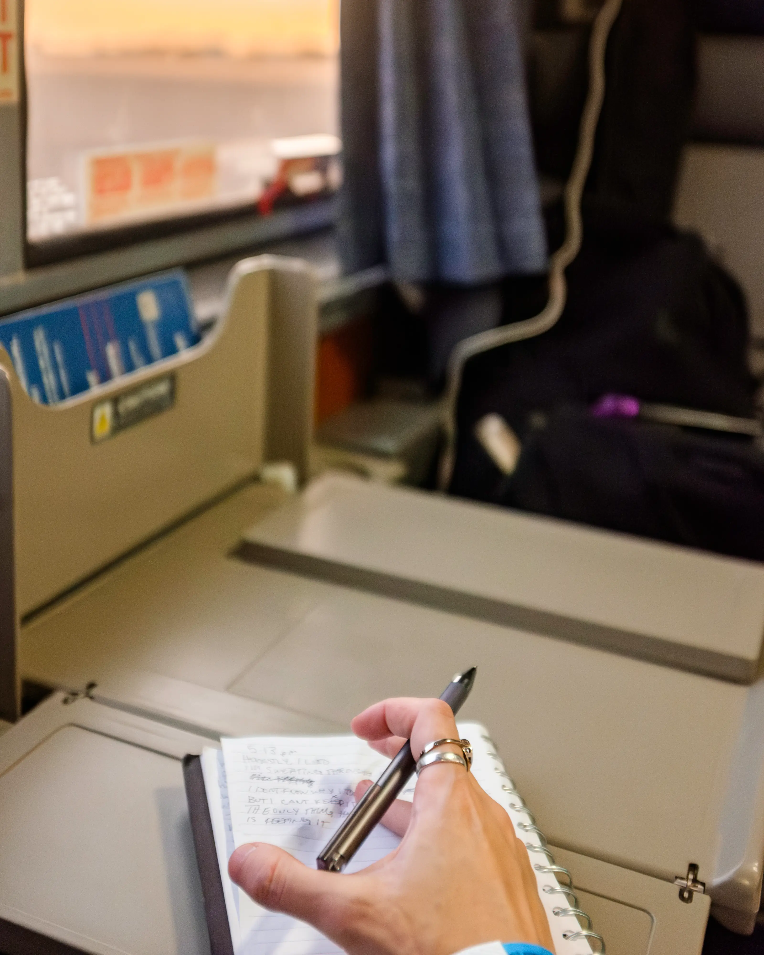 A hand holding a pen above a notebook on a train table in front of a window