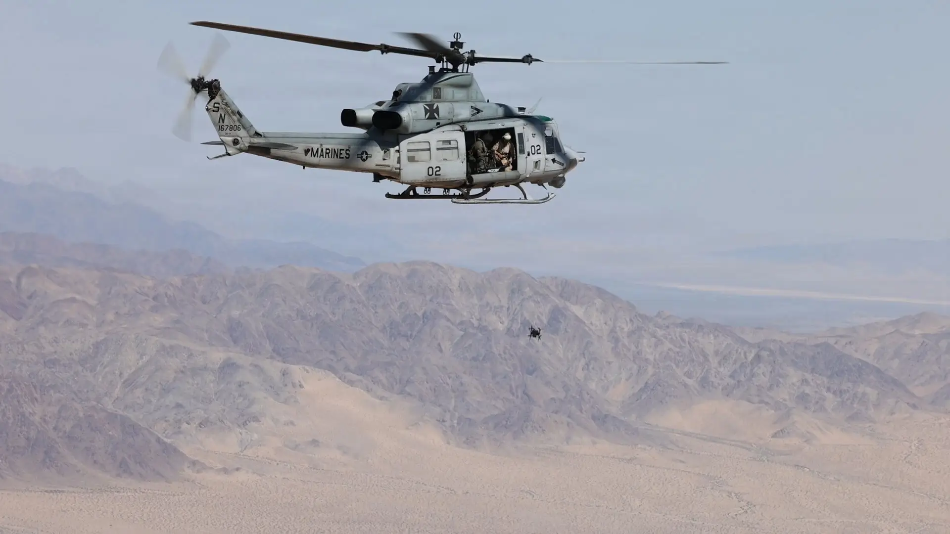 A UH-1Y Venom helicopter flies above a sandy, rocky terrain with a small drone below it.