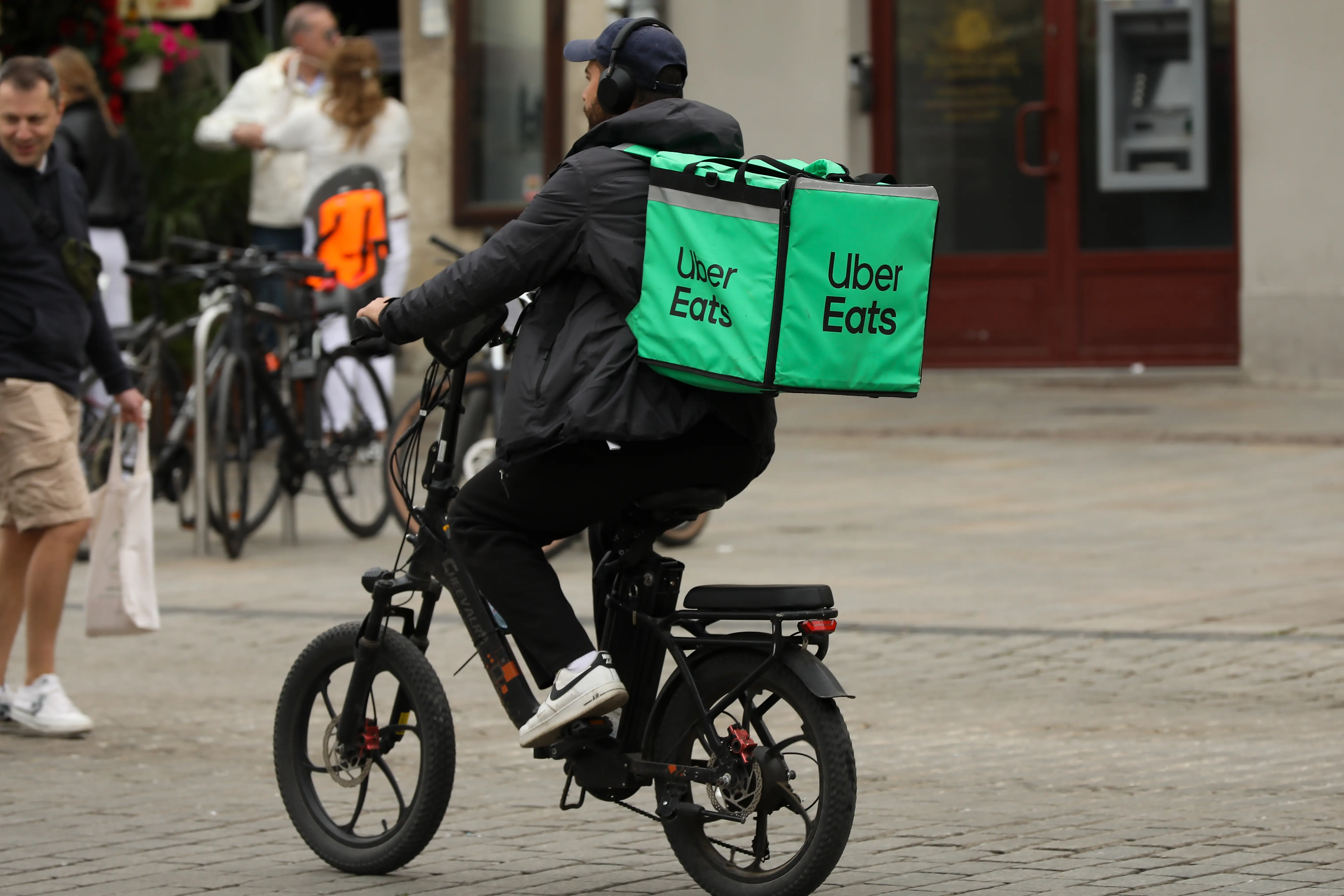 A bike courier wears an all-black outfit and headphones along with a light-green Uber Eats backpack as he rides through the center of a city.