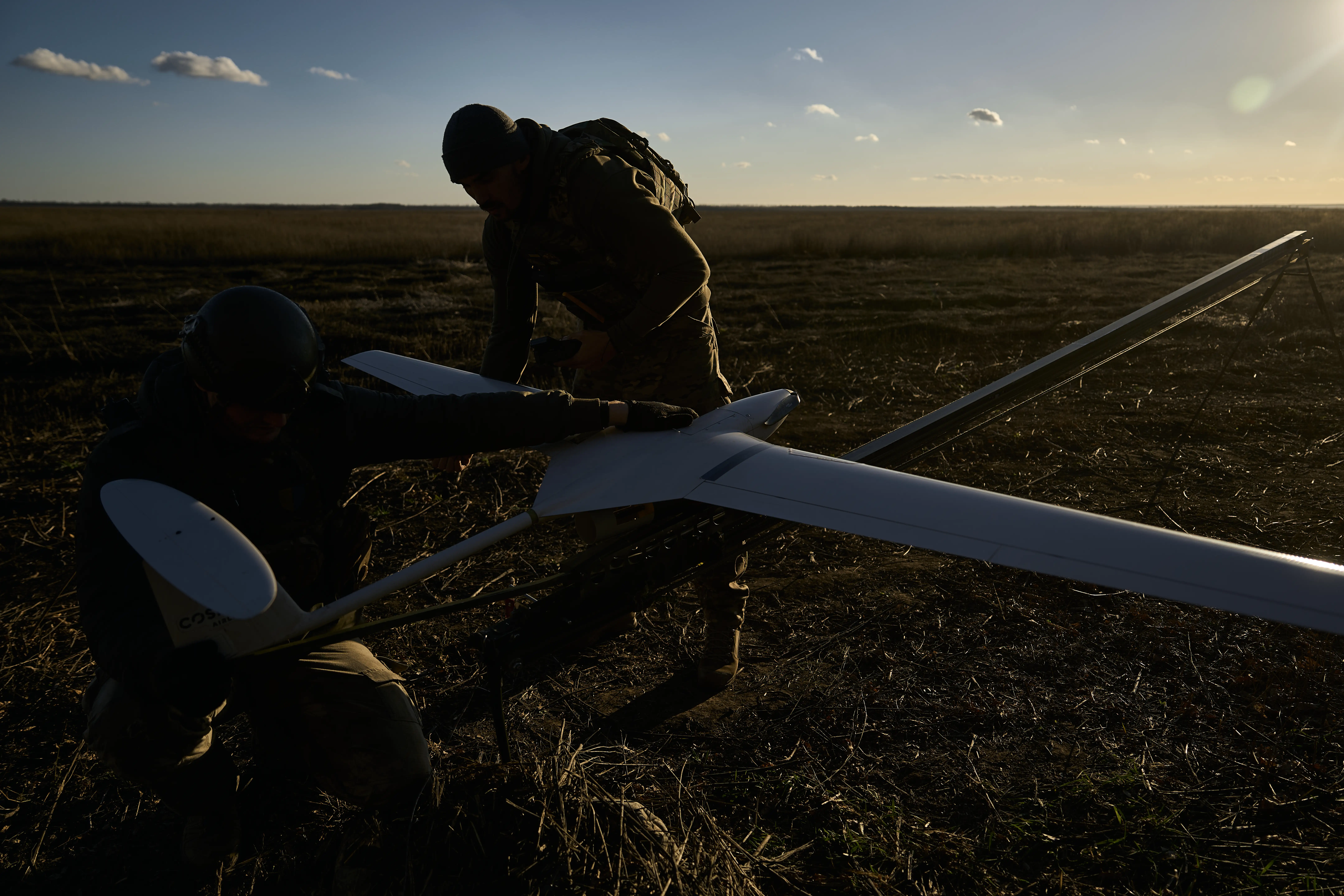A man silhouetted in the sun bends over a large grey drone in a field