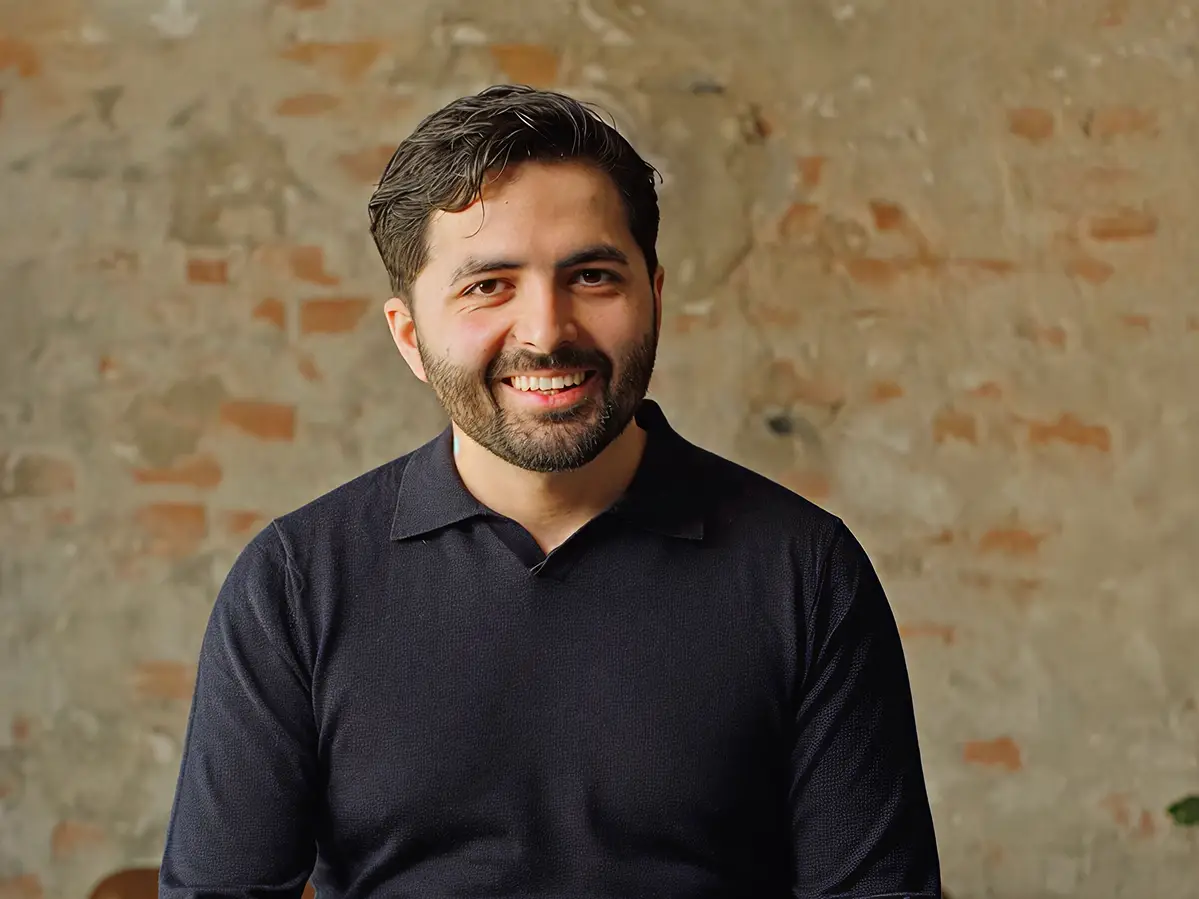 A smiling man in a dark polo shirt sits in front of an exposed brick wall and leafy plant.