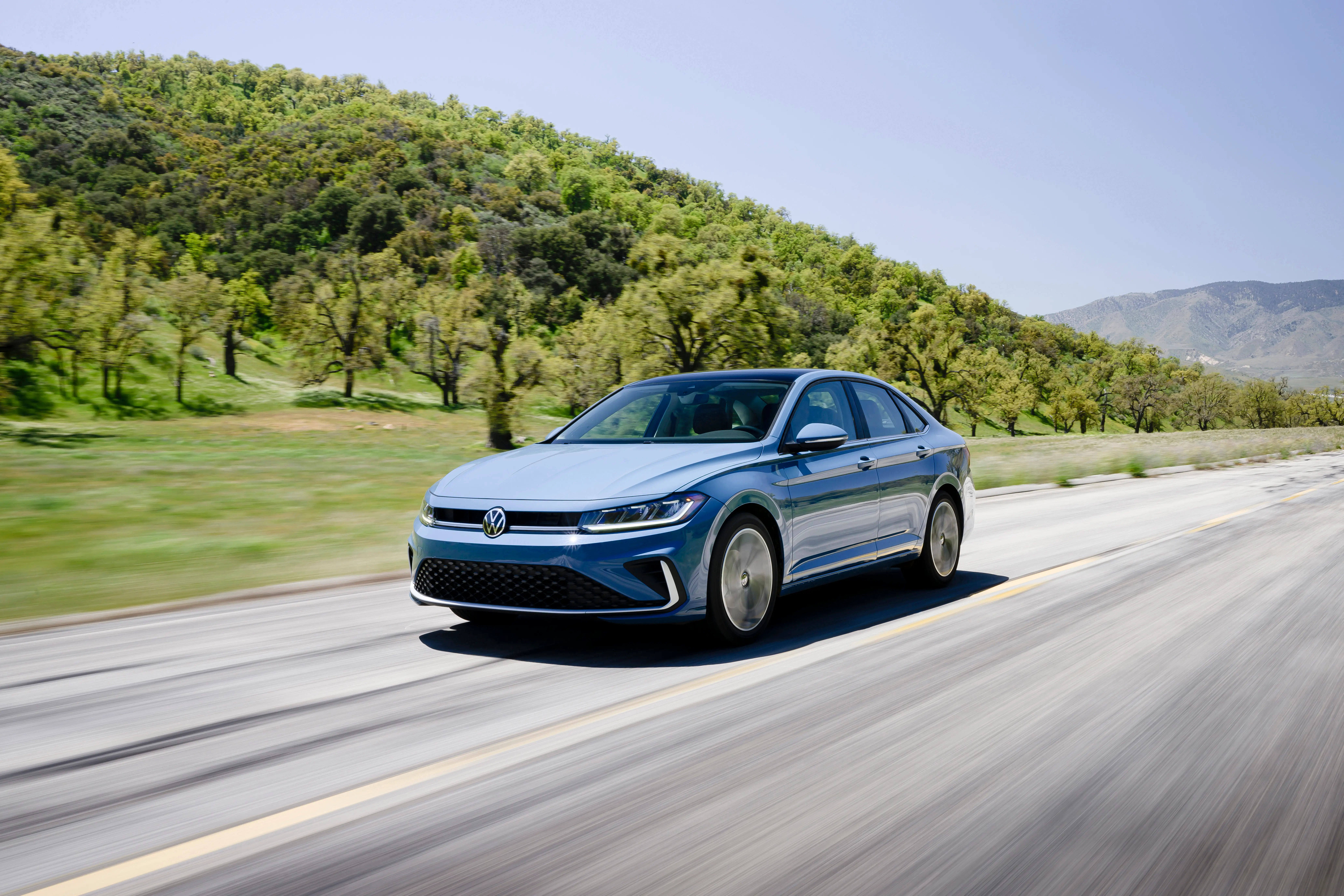 A light blue Volkswagen Jetta sedan drives down a two-lane road past a hill with trees.