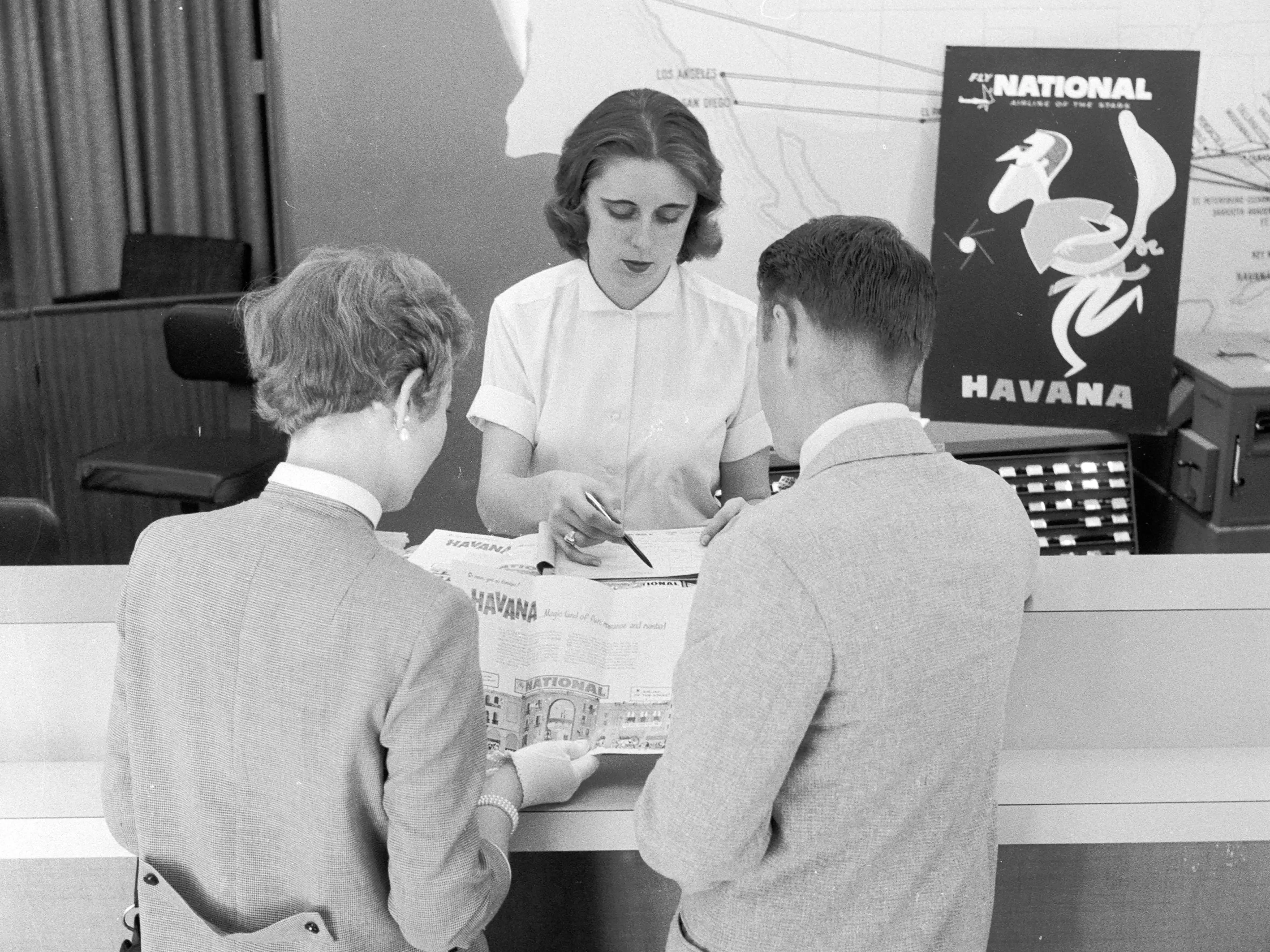 A couple, a man and a woman, look over a brochure touting a package vacation to Havana, Cuba, for National Airlines while airline staff point out details in the brochure, May 1957.