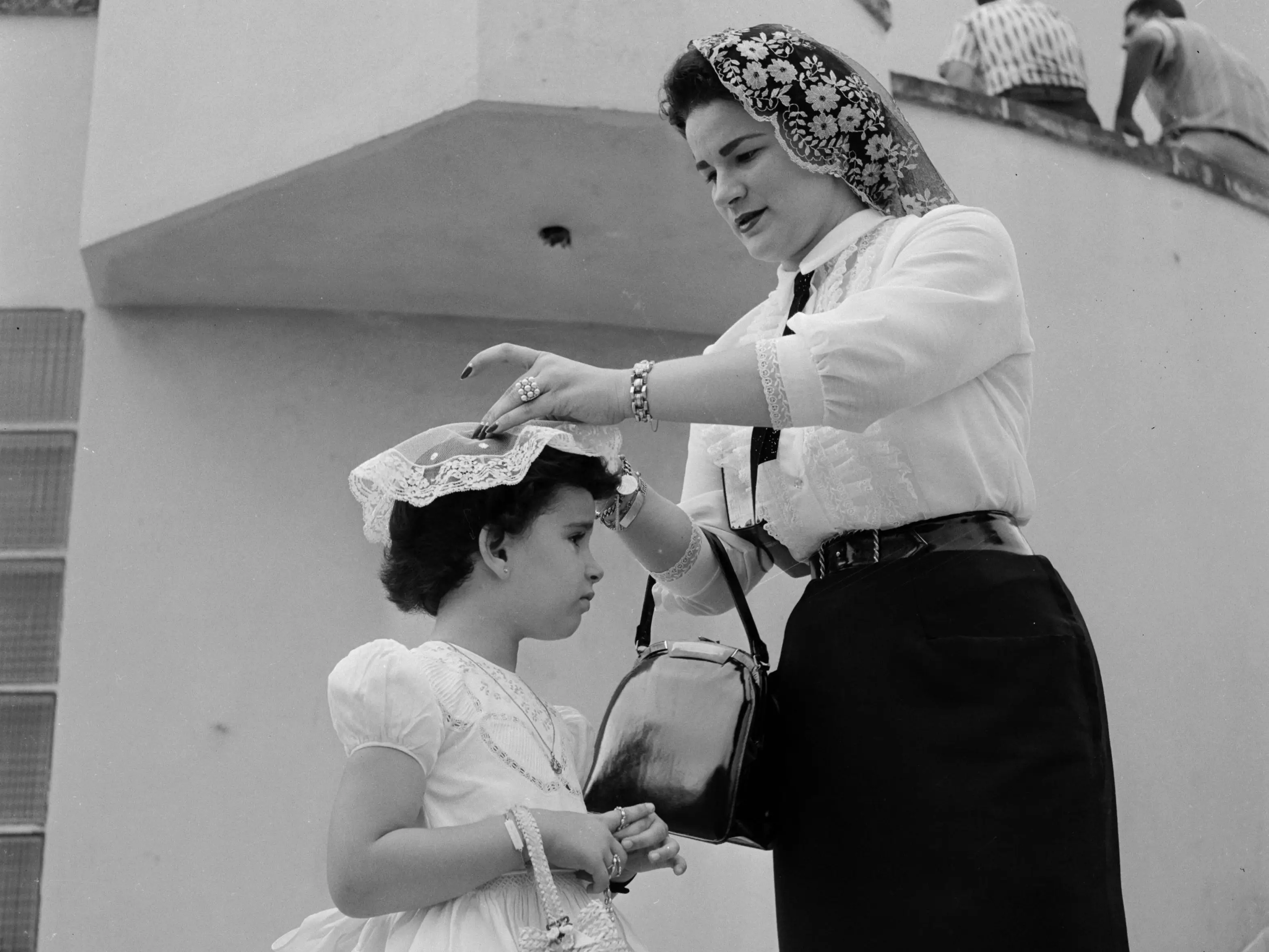 circa 1955: Delia Gondar fixes a customary 'velo' headpiece on to her daughter Delia Maria's hair before they go into church for Sunday mass at Marianao, Cuba.