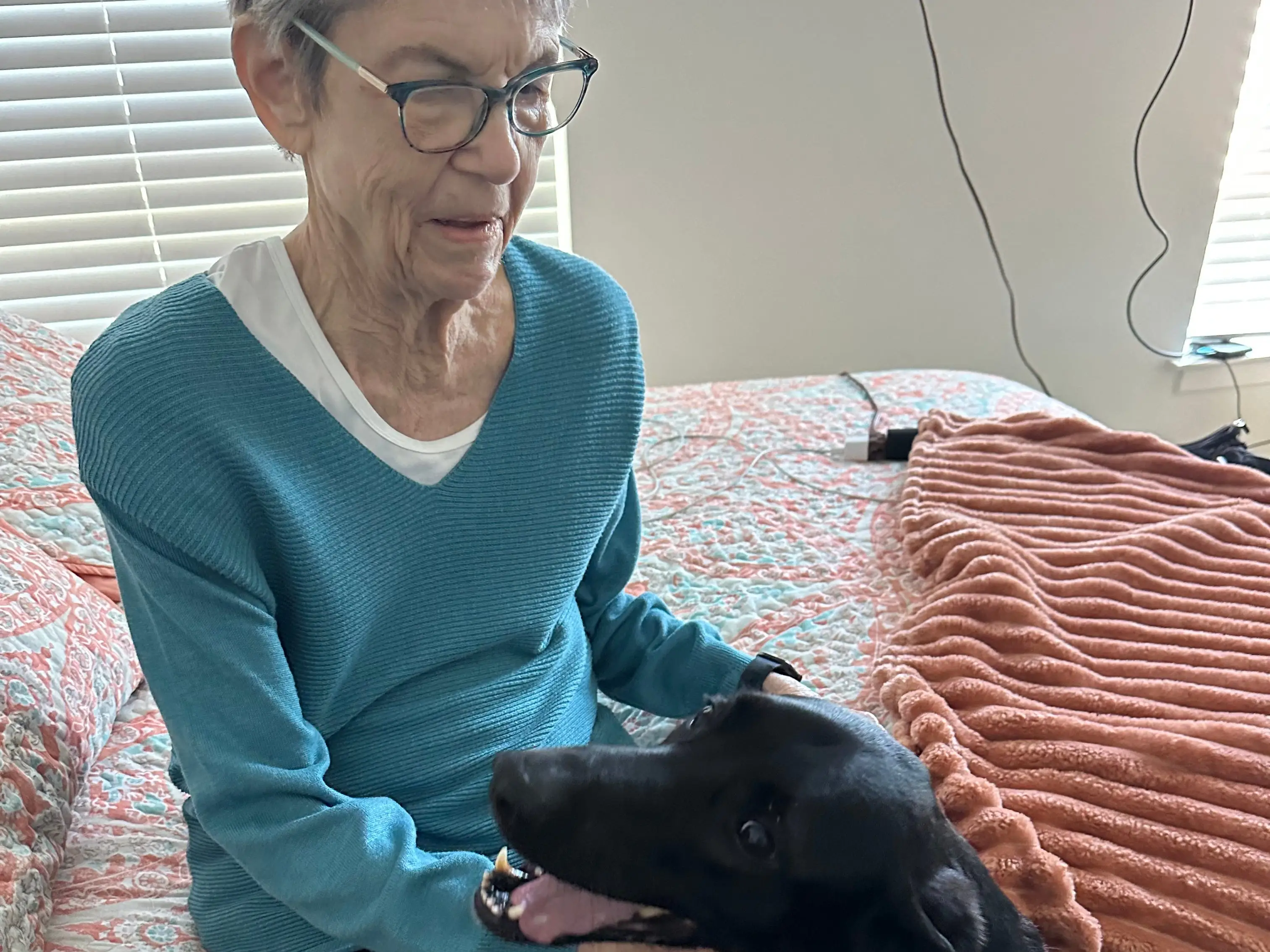 An older woman sitting on a bed petting a black dog.