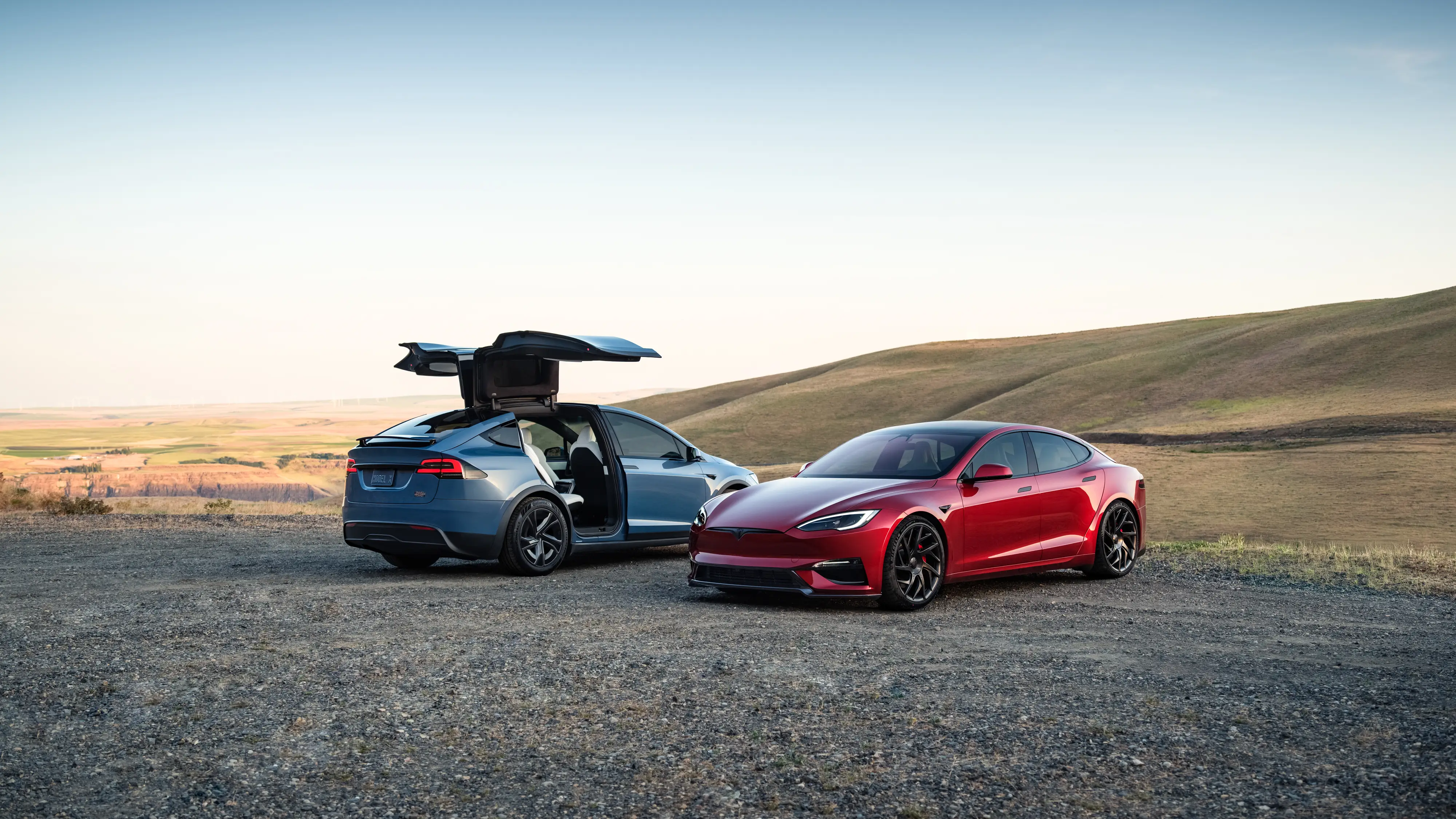 A blue Tesla Model X, with its rear doors open, is parked next to a red Tesla Model S on a gravel lot.