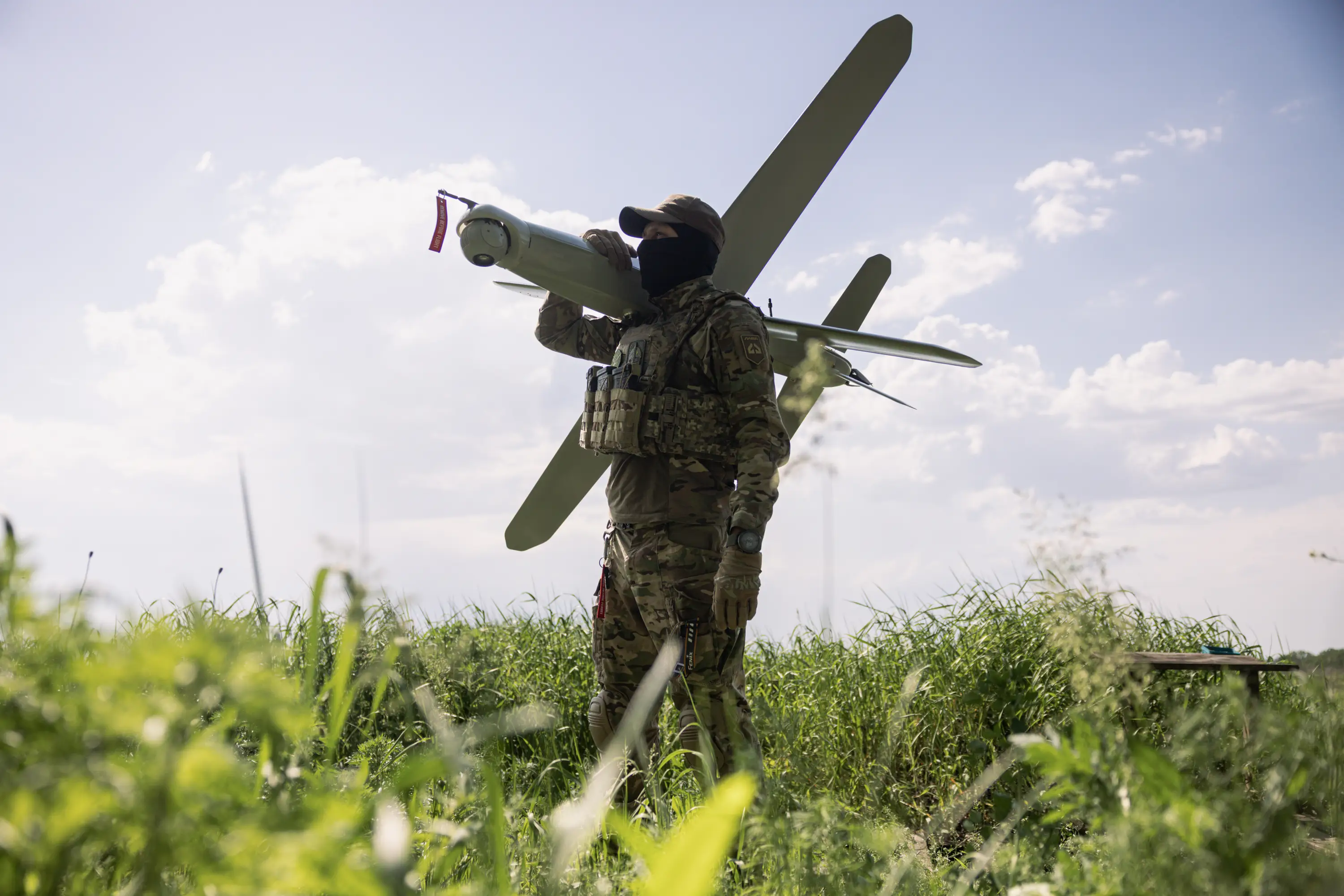 A man in camouflage and a black balaclava stands in a sunny field with a large grey drone over his shoulder