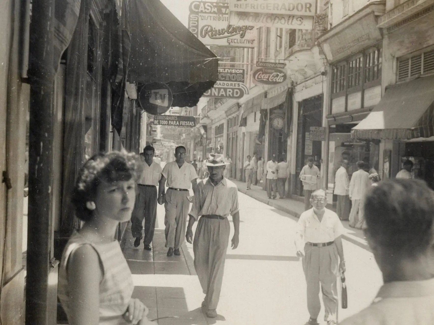 View of a young woman in a sleeveless dress and a flower headpiece, her purse and sunglasses in her hands, as she stands in the shade on an unidentified, shop-lined street, Cuba, mid 1955.