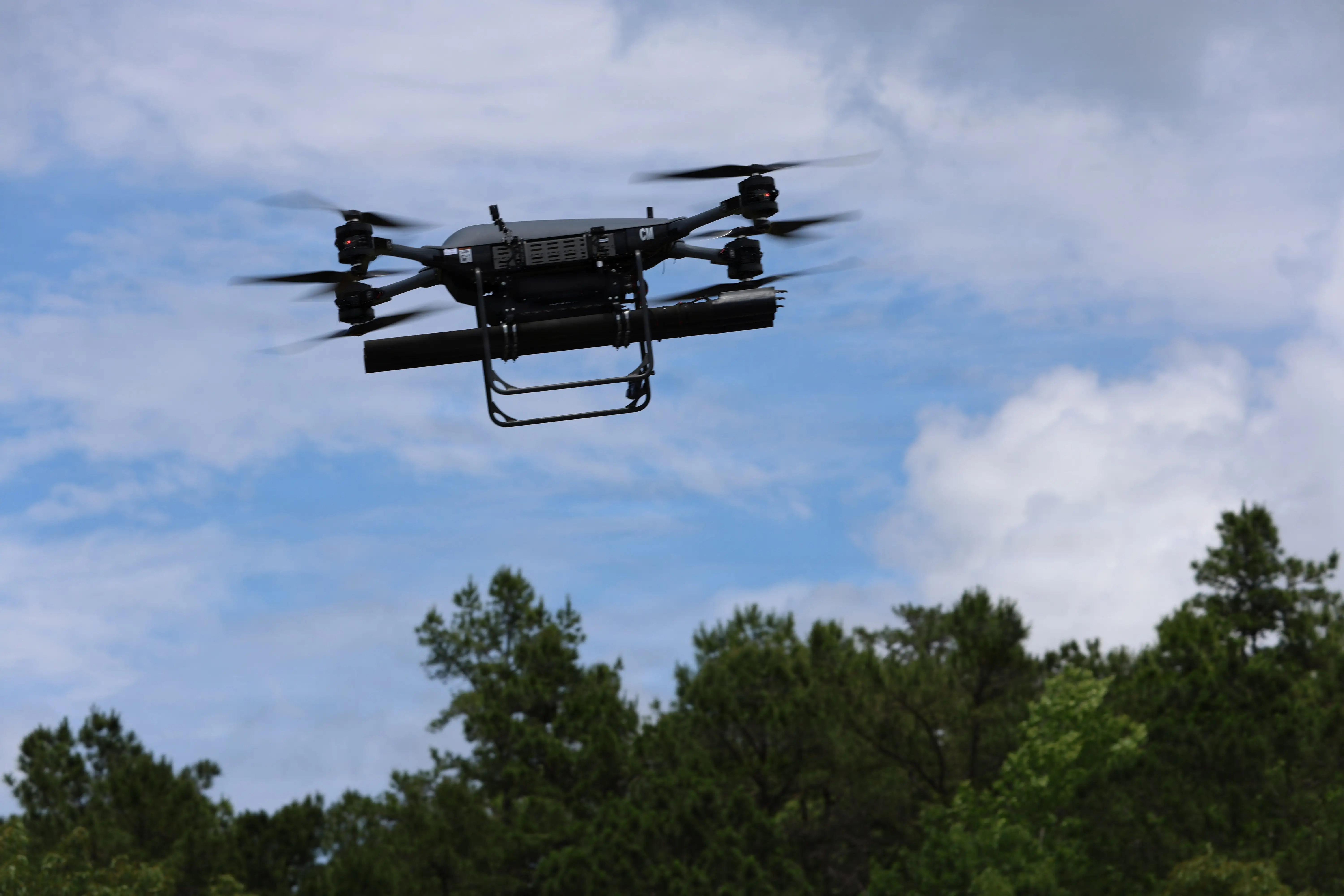 A black drone flying in the sky above a line of trees.