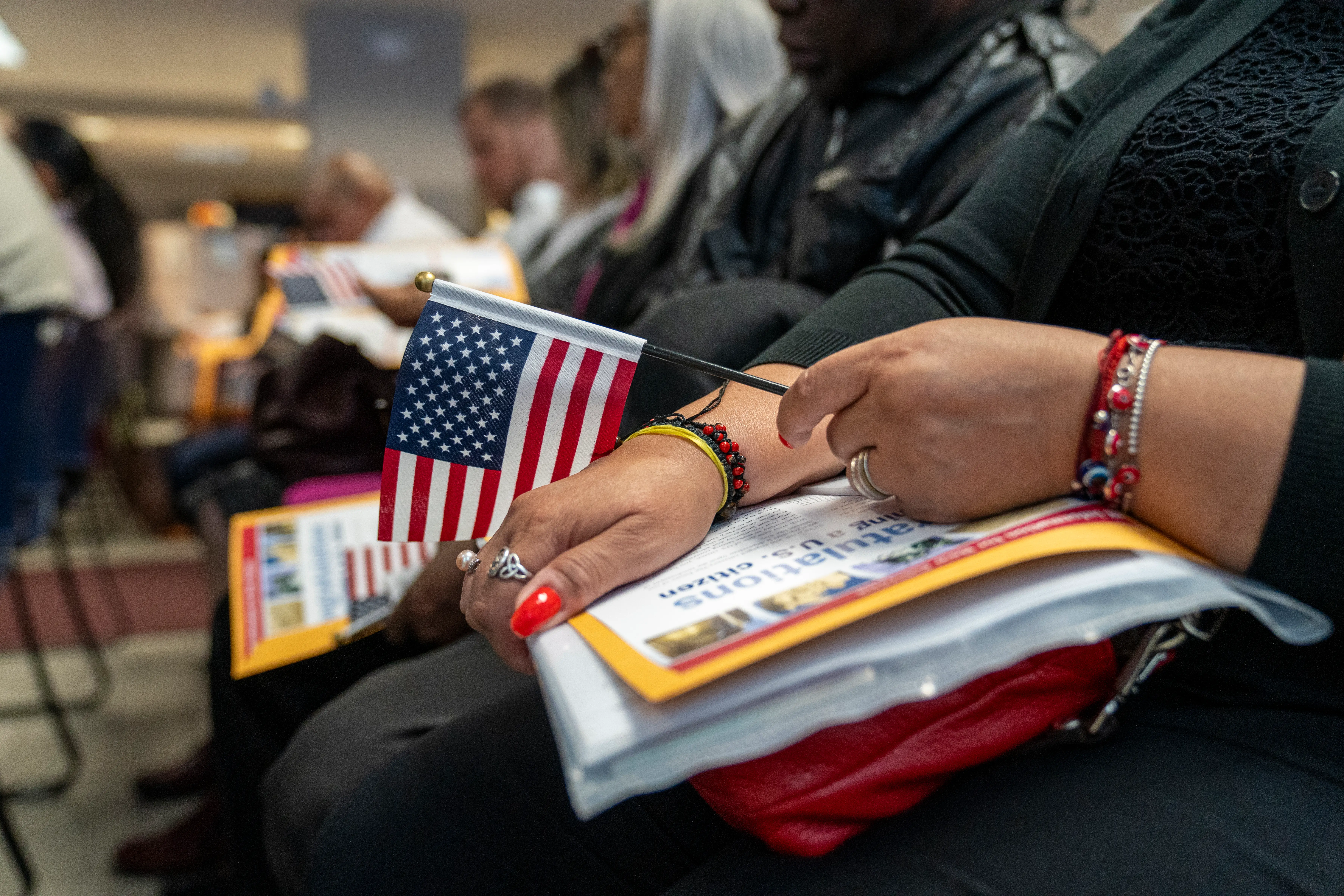 A row of candidates for US citizenship sits in chairs. The closest is holding an American flag.