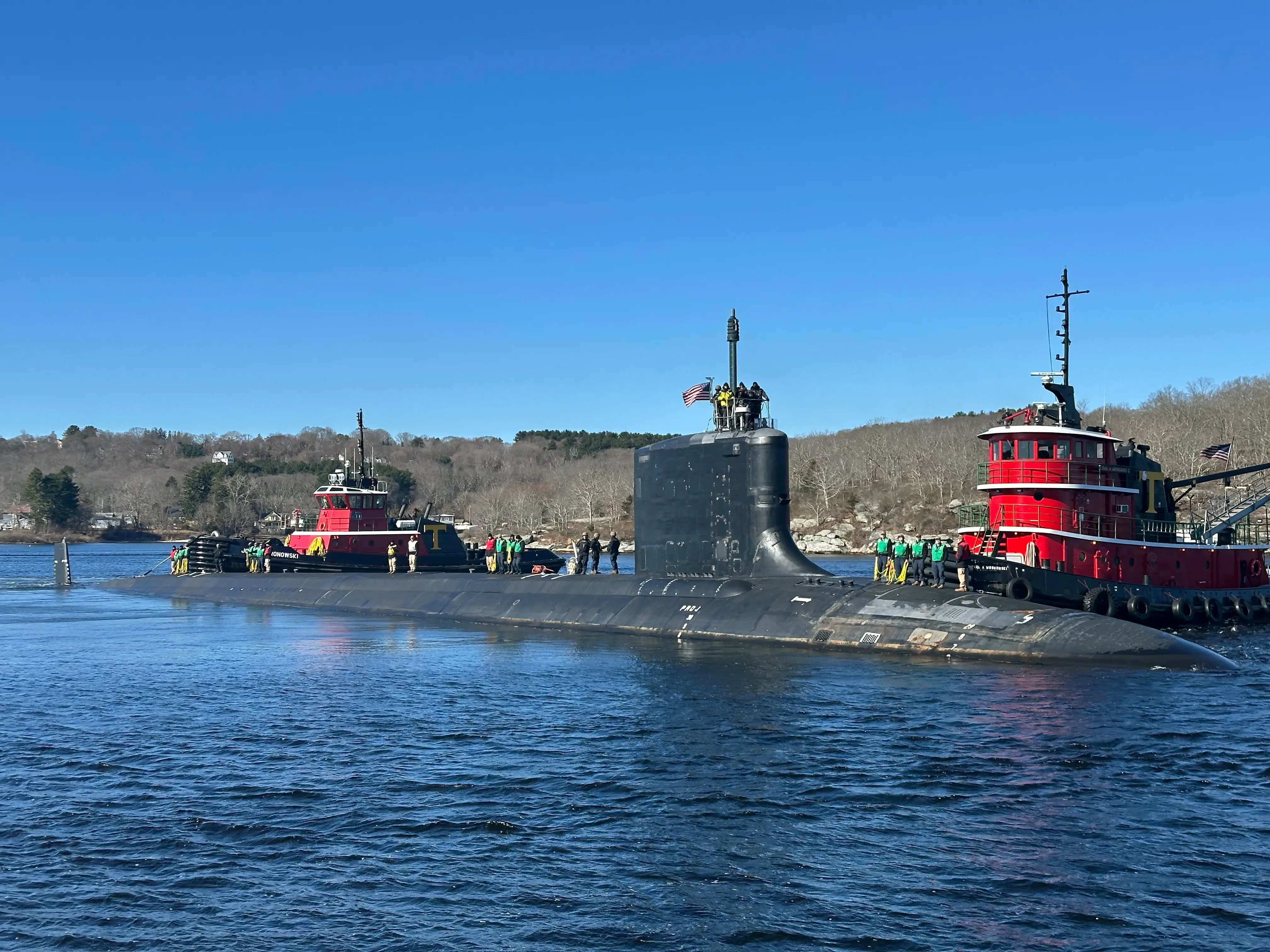 A submarine sits in blue water near a coast. People stand on the submarine.