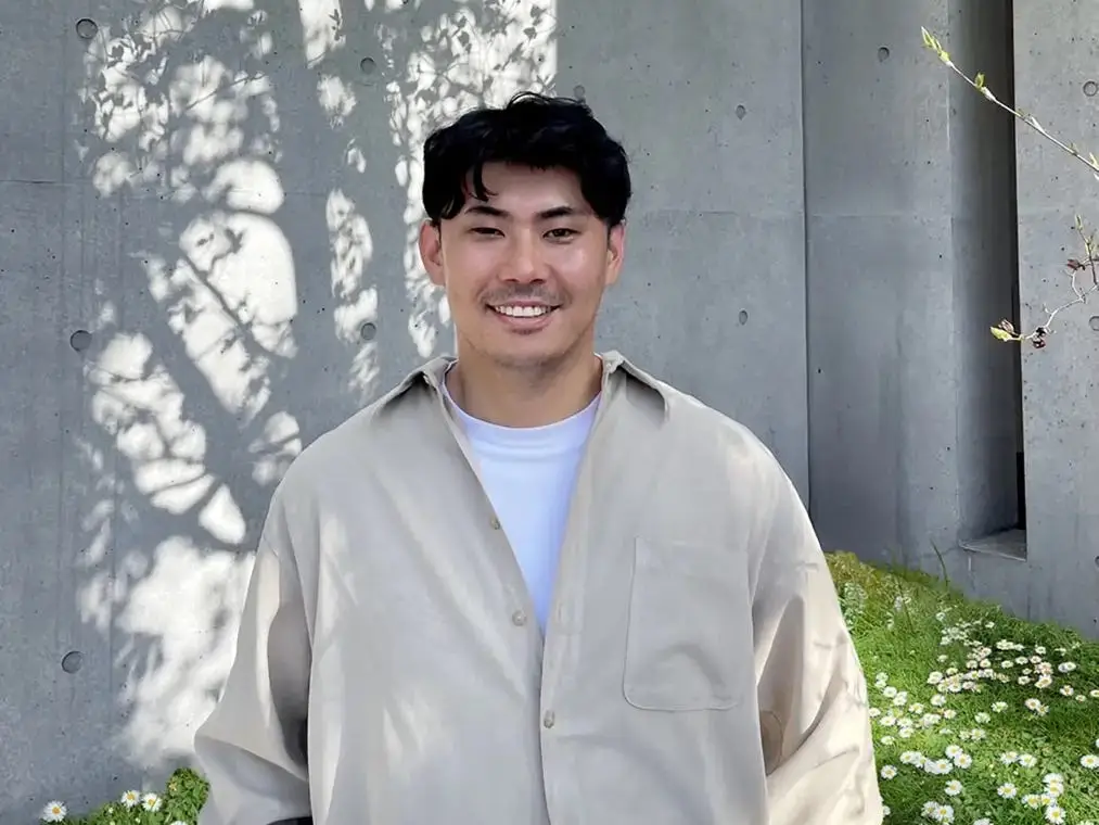A man wearing a beige shirt standing in front of a concrete wall.