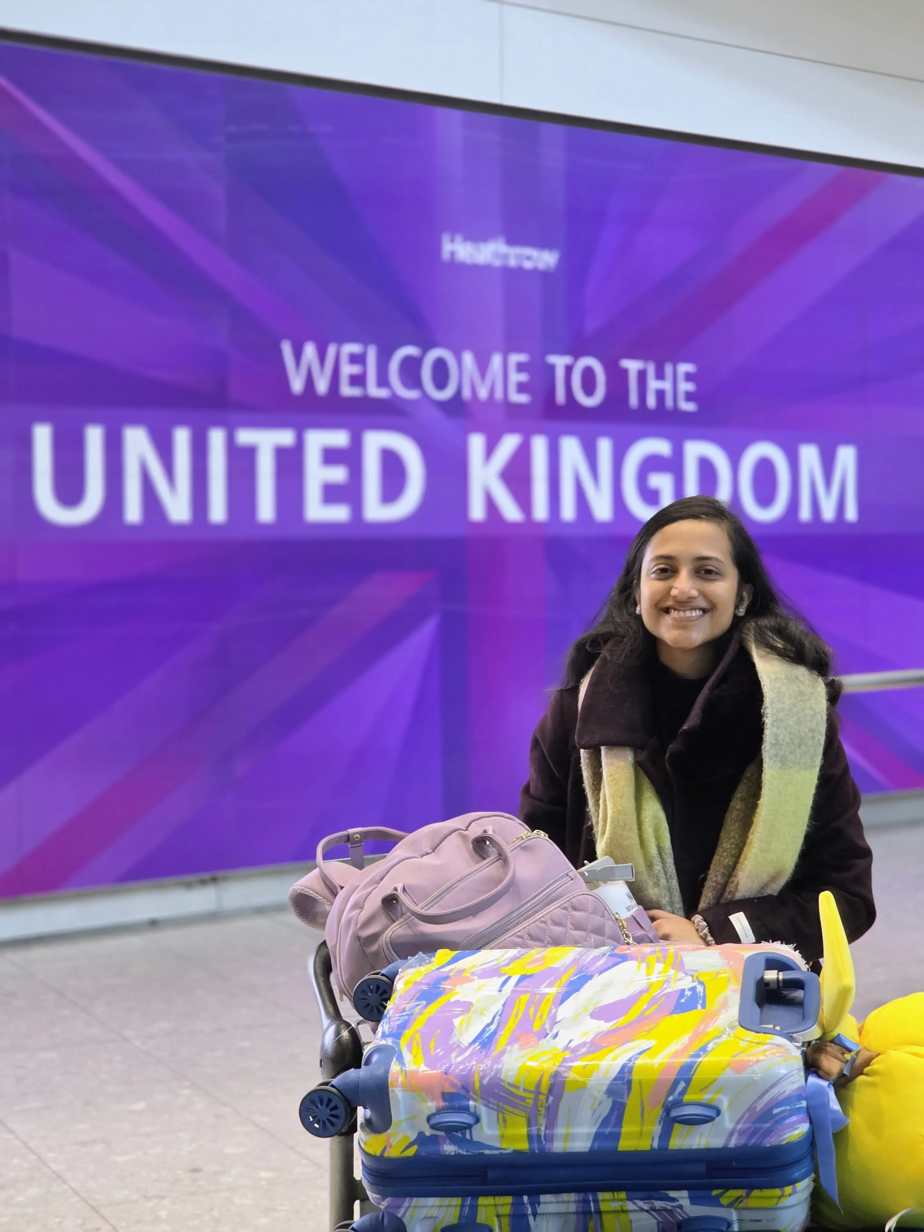 Sunjana Ramana is standing in Heathrow Airport in front of a sign that says 