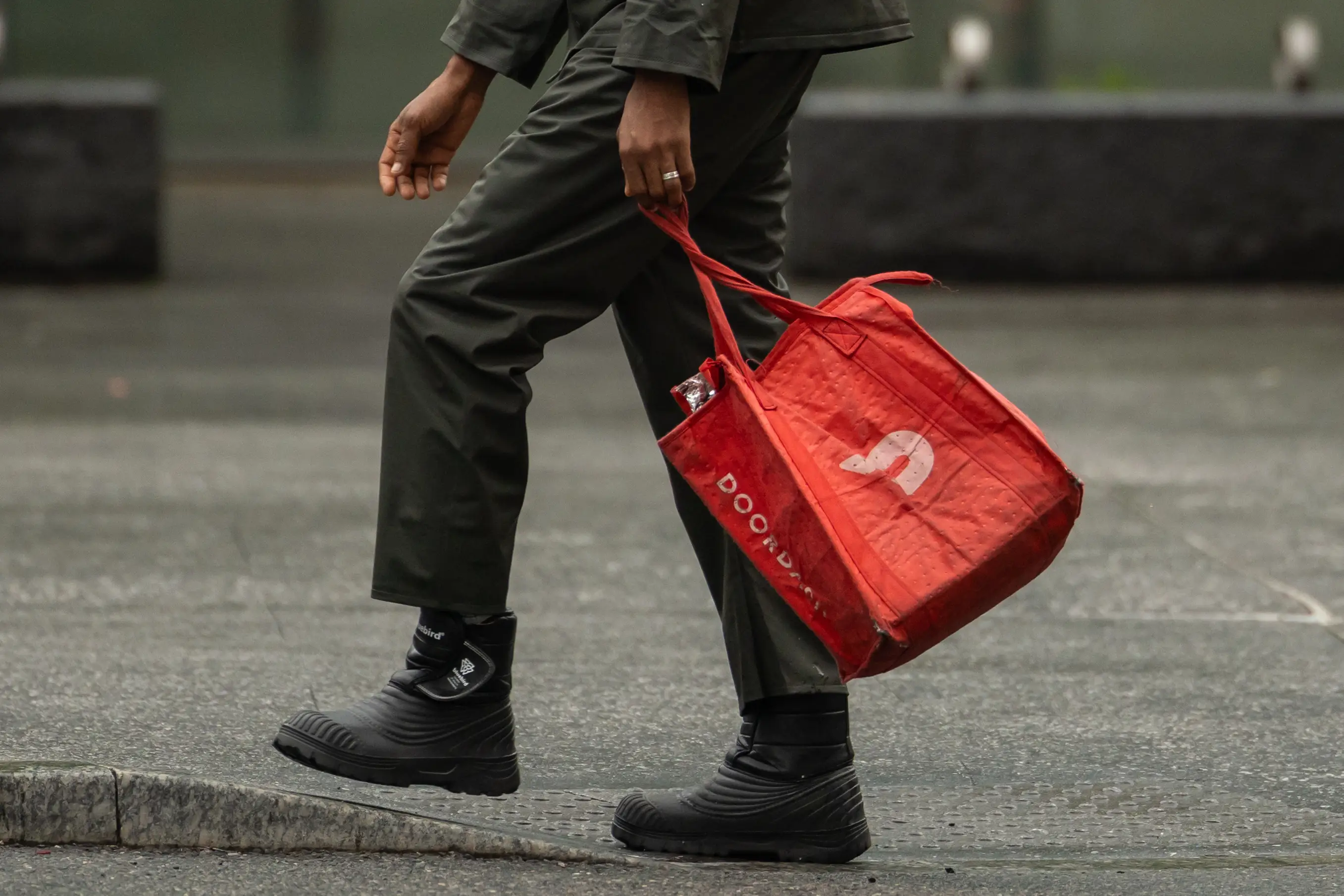 A pedestrian walks on a sidewalk while carrying a red DoorDash-branded insulated bag.