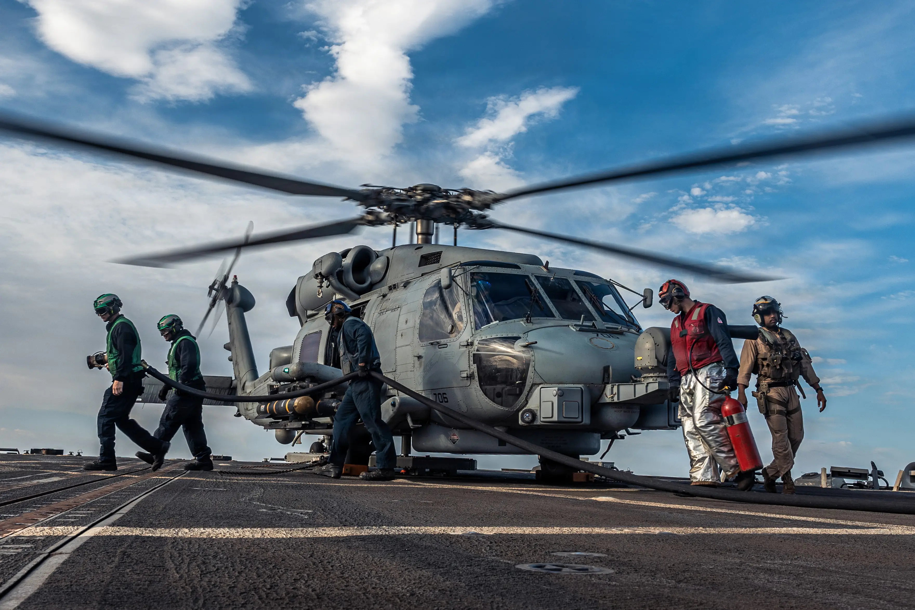 A group of military personnel walk around a helicopter on the deck of a warship.
