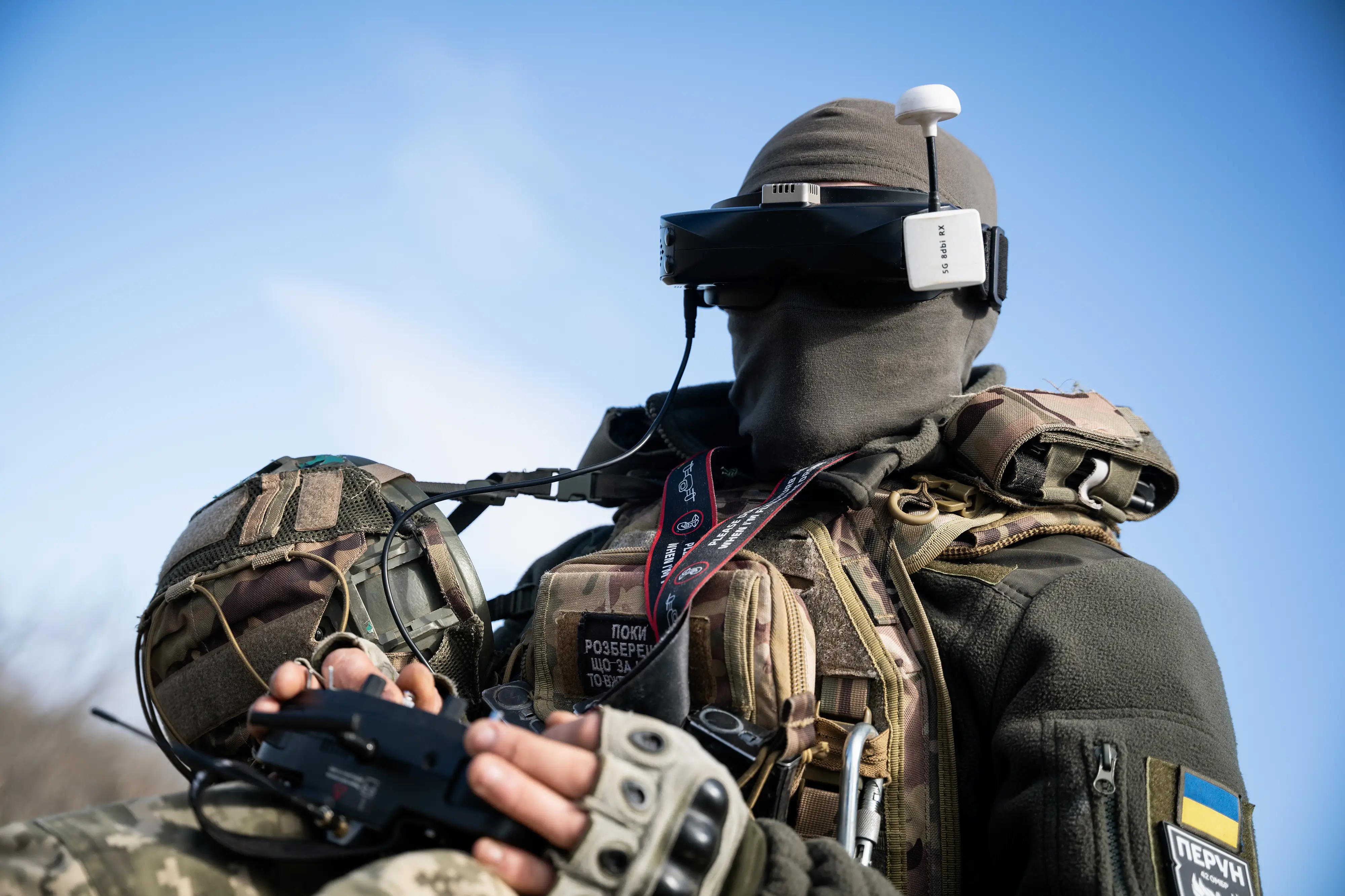 A man in khaki and camouflage clothing sits under a blue sky, holding a controller, wearing a balaclava and goggles