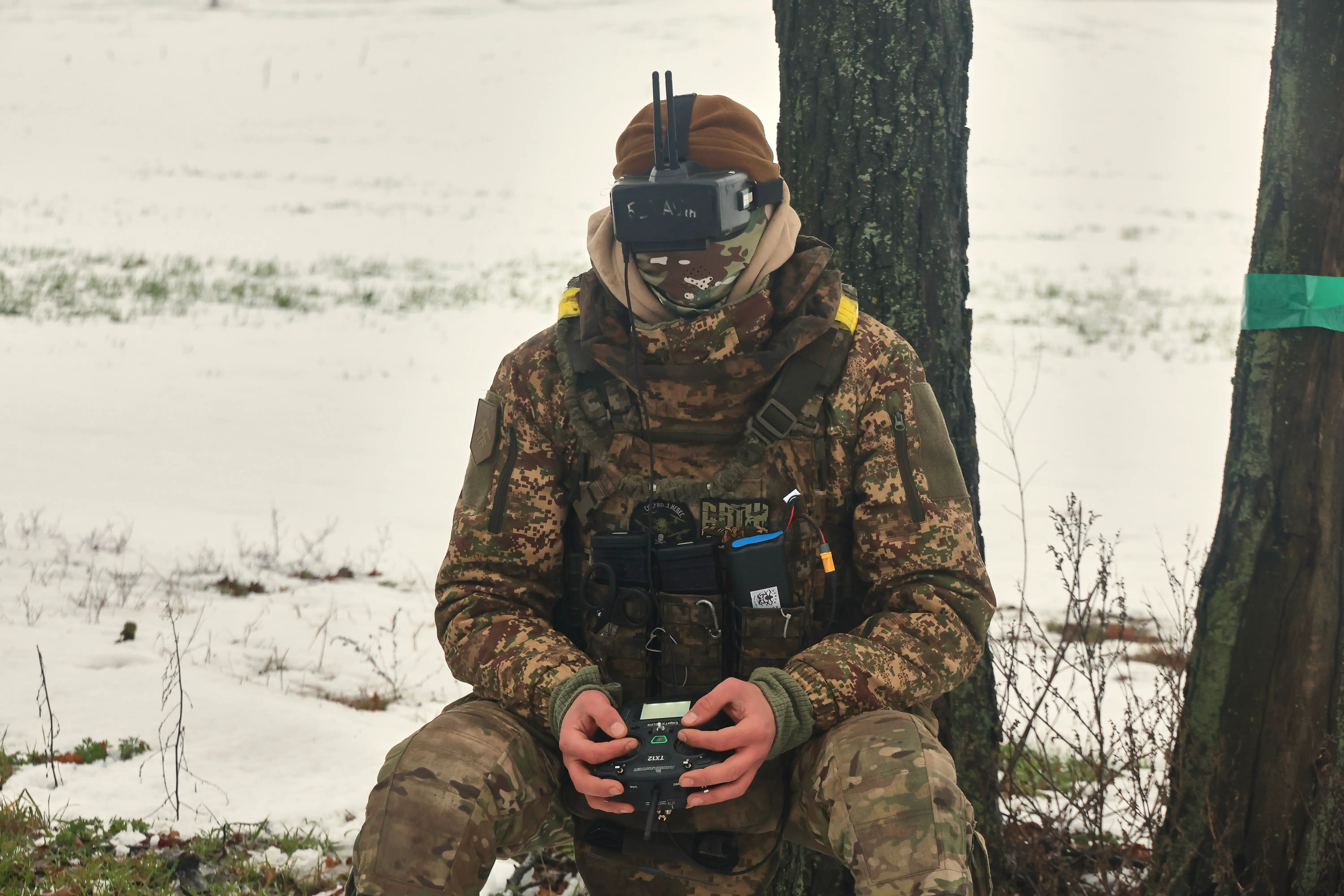 A man in camouflage gear and a beanie hat sits beside a tree in the snow holding a controller and with a headset over his eyes