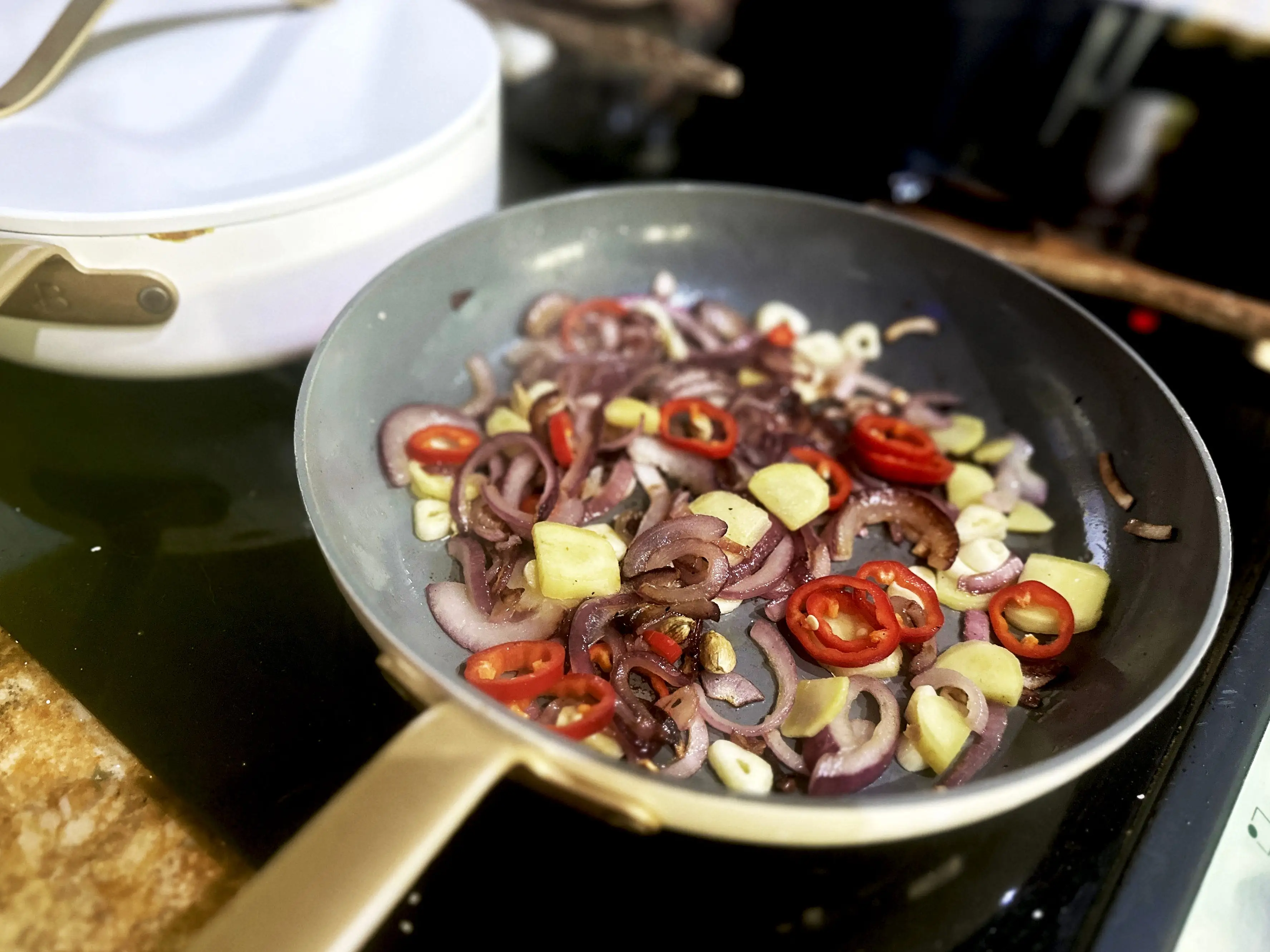 A pan filled with cooking chile peppers and sliced red onions next to a white pot with a lid on it
