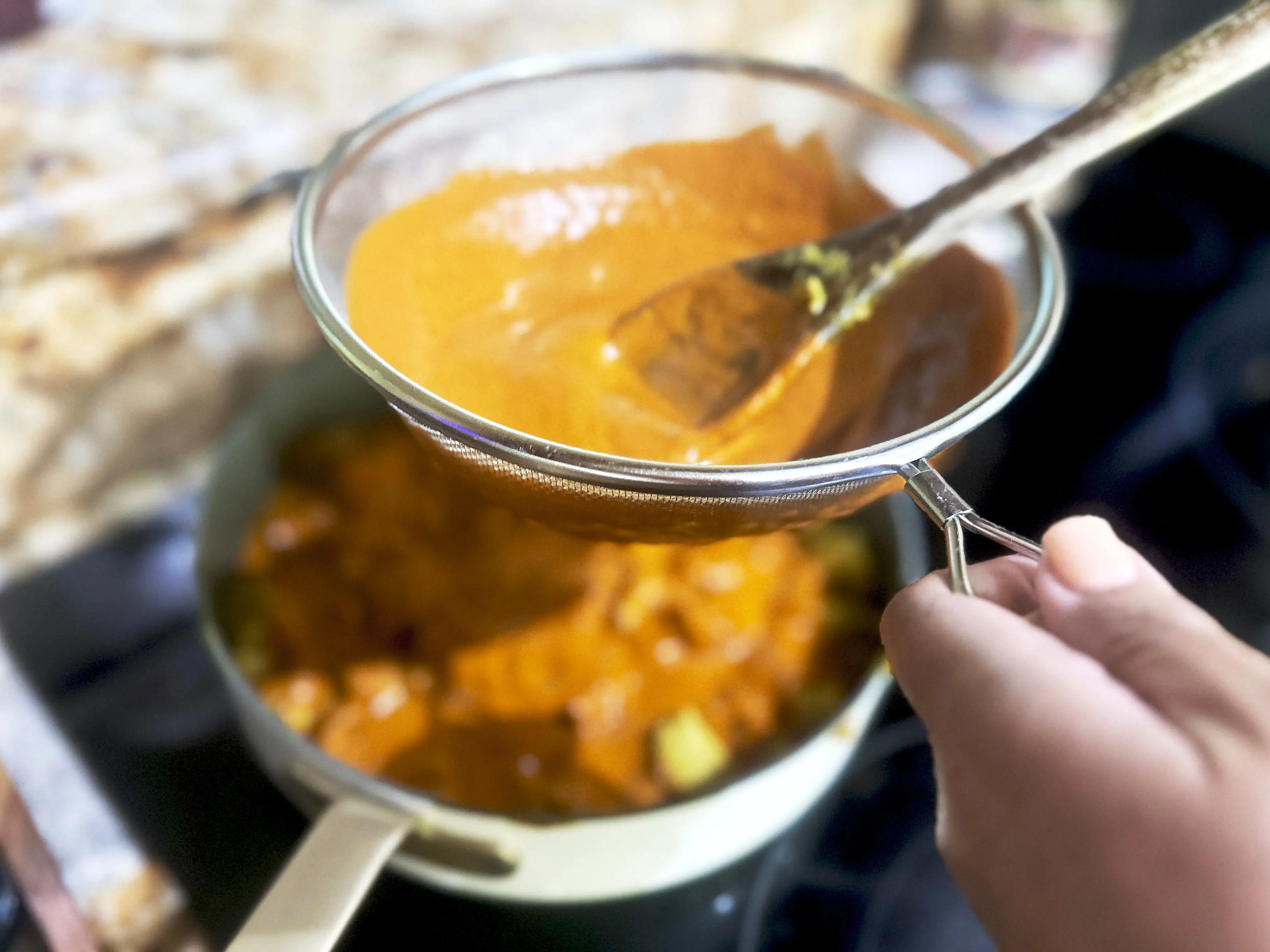 An orange sauce being strained through a sieve into a larger pan. A black stove range sits in the background