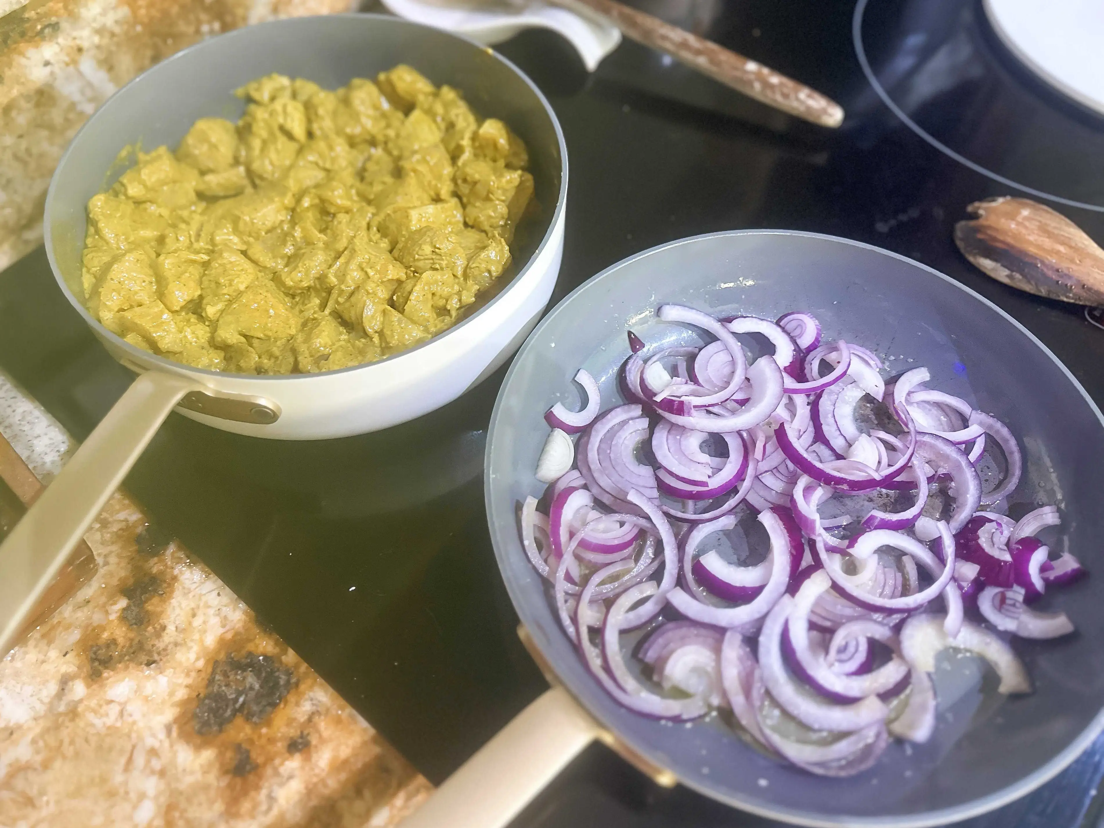 Two pans sitting next to each other on a black stovetop range. One pan holds chicken pieces in a yellow-brown marinade and the other holds slices of red onion