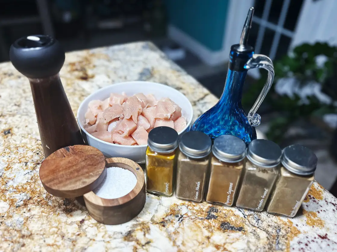 A cup of salt, a pepper grinder, a white bowl of raw chicken pieces, an olive-oil dispenser, and several spices lined up on a marble kitchen counter