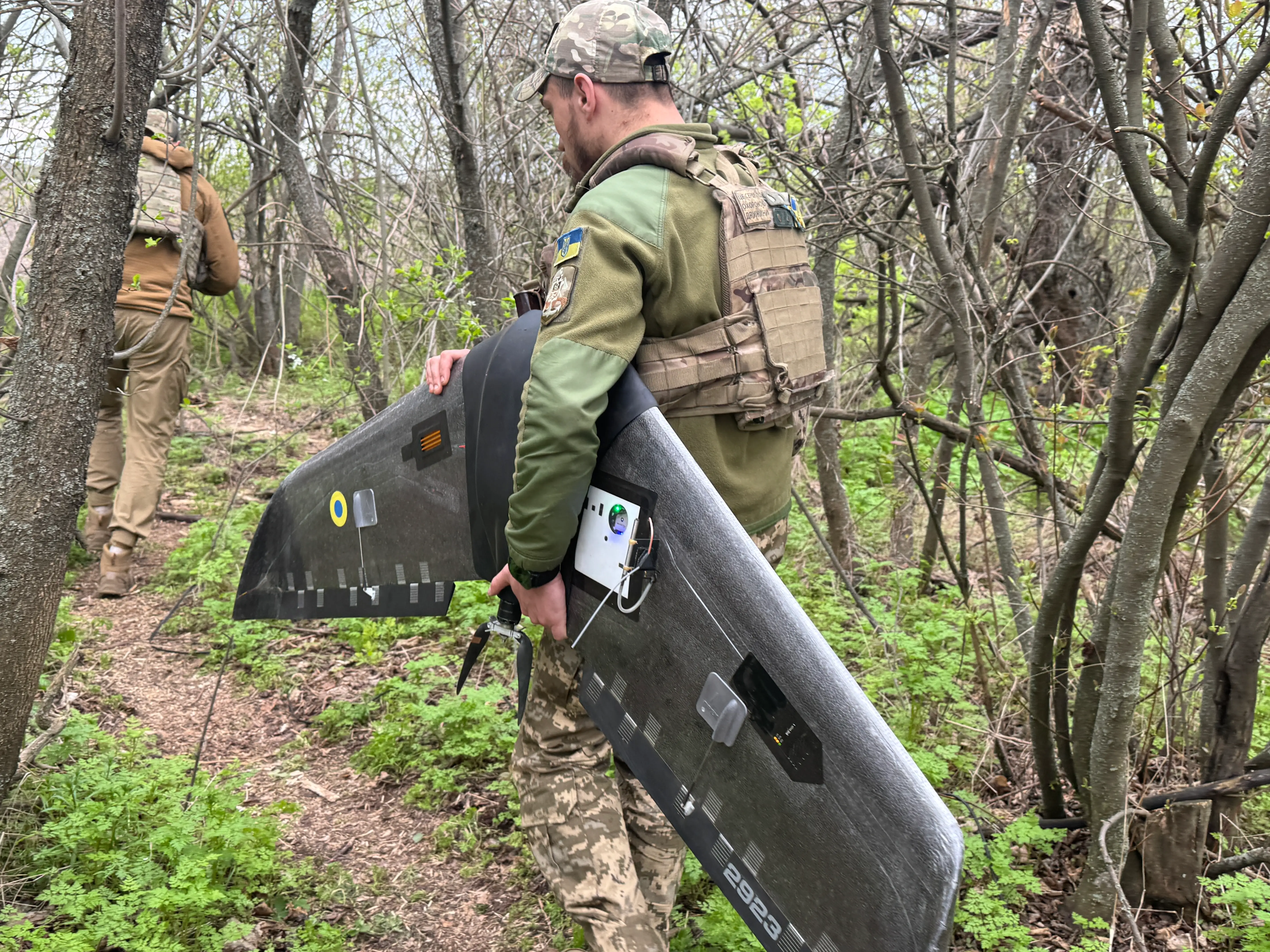 A man in khaki carring a large black drone under his arm between trees
