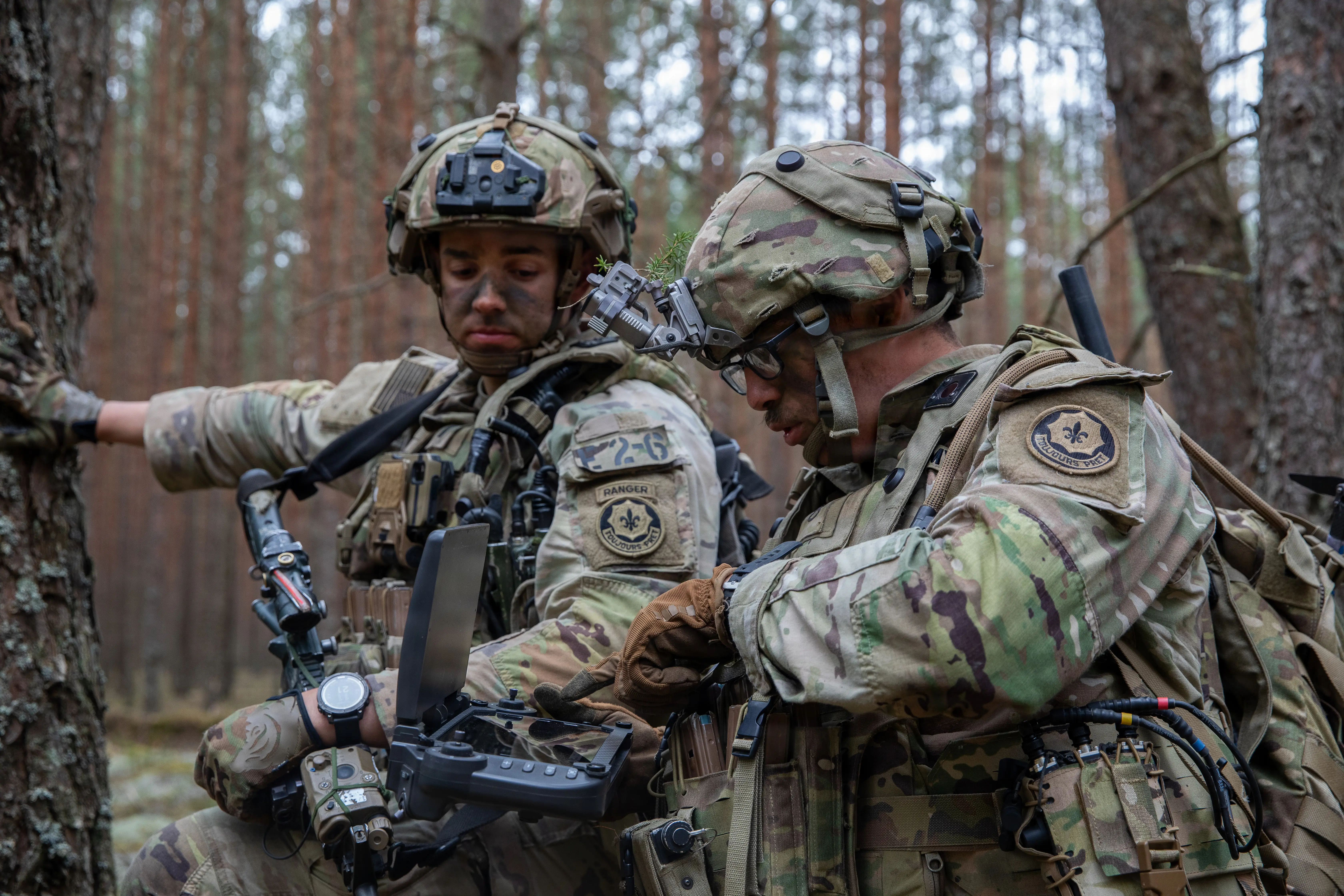 Two US soldiers stand amongst trees looking at a drone controller.