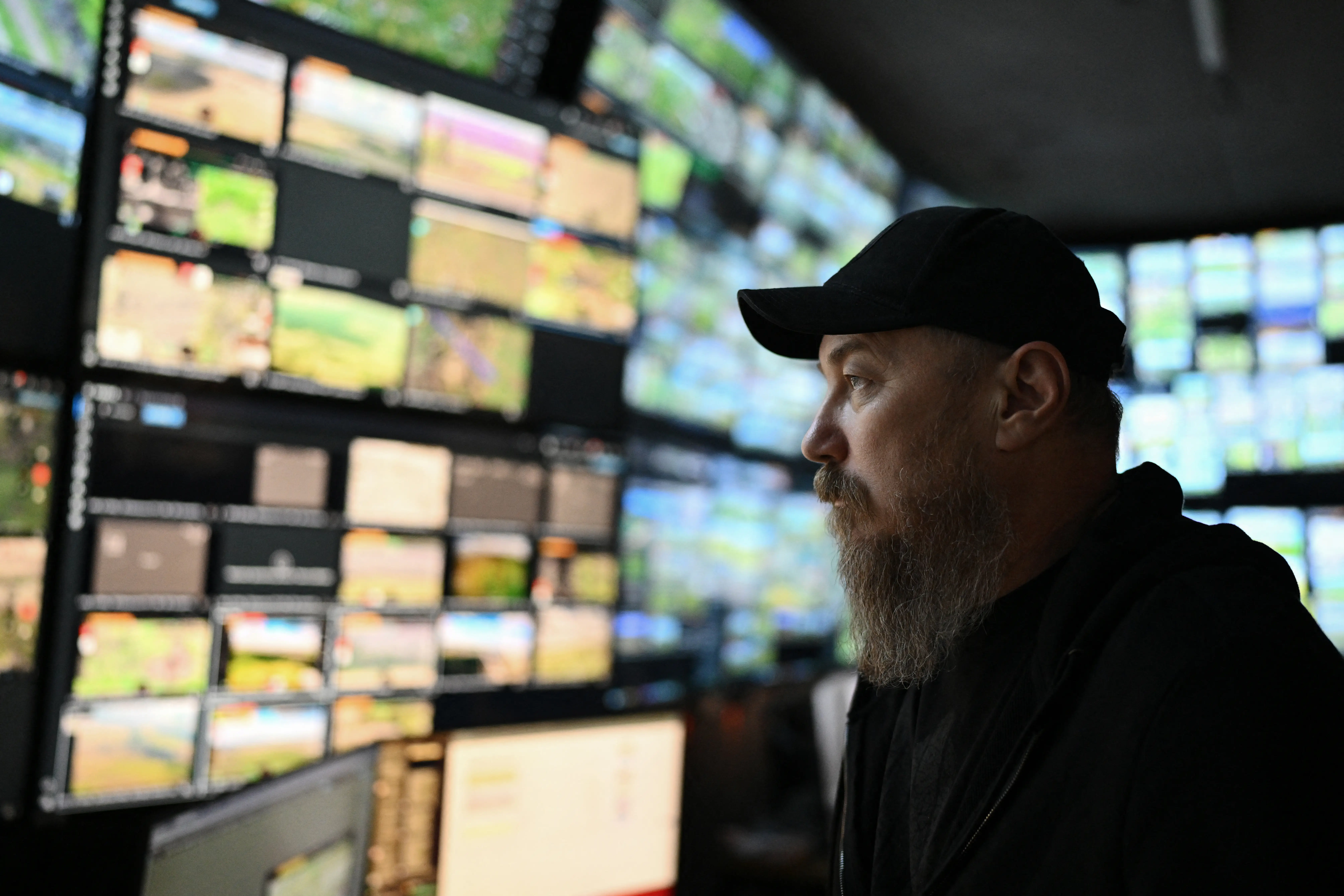 A man in a black cap and a beard looks at a wall of data screens in an indoor location